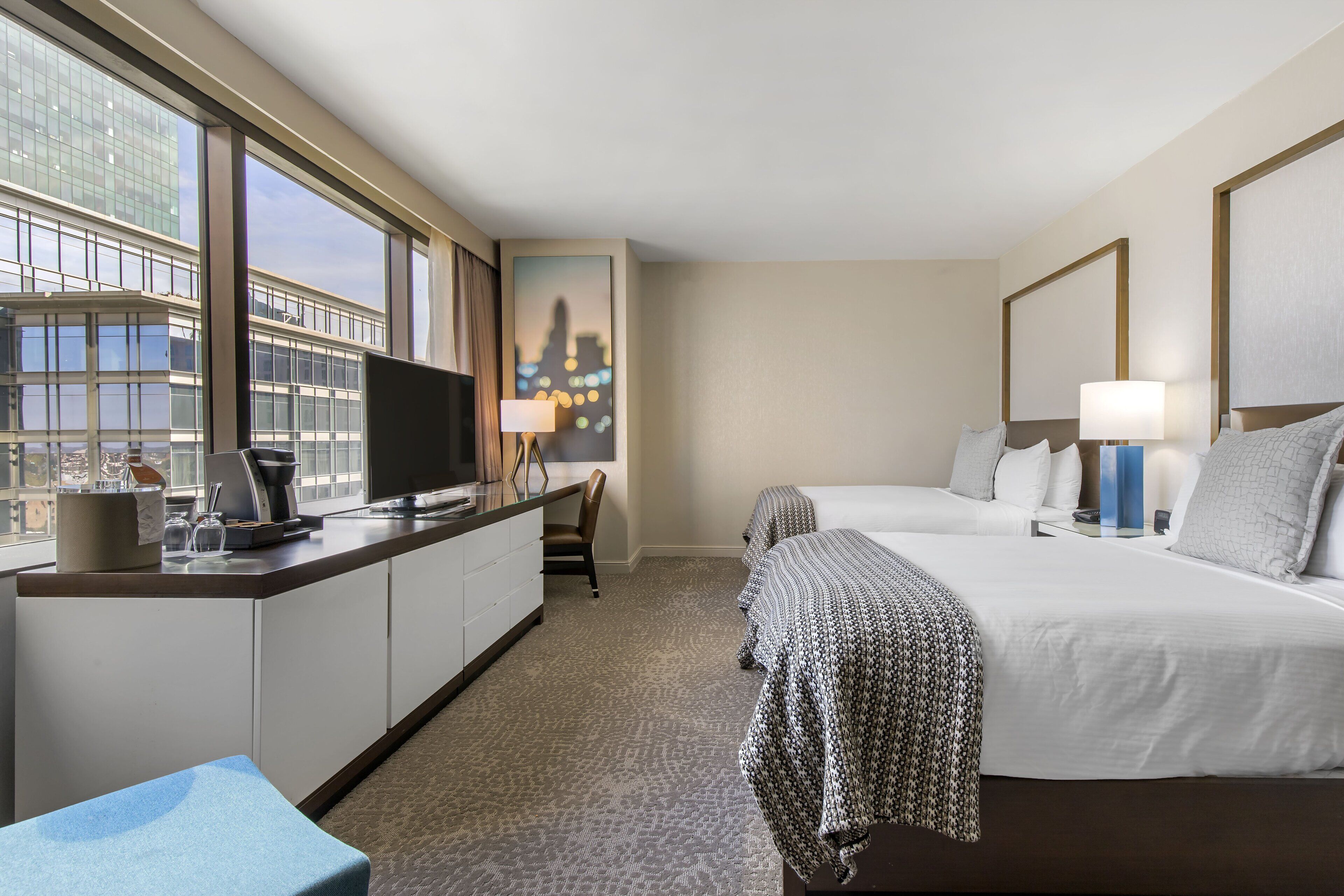 A hotel room with a large window, a desk with a computer and coffee maker, two beds with white bedding, and a cityscape view.