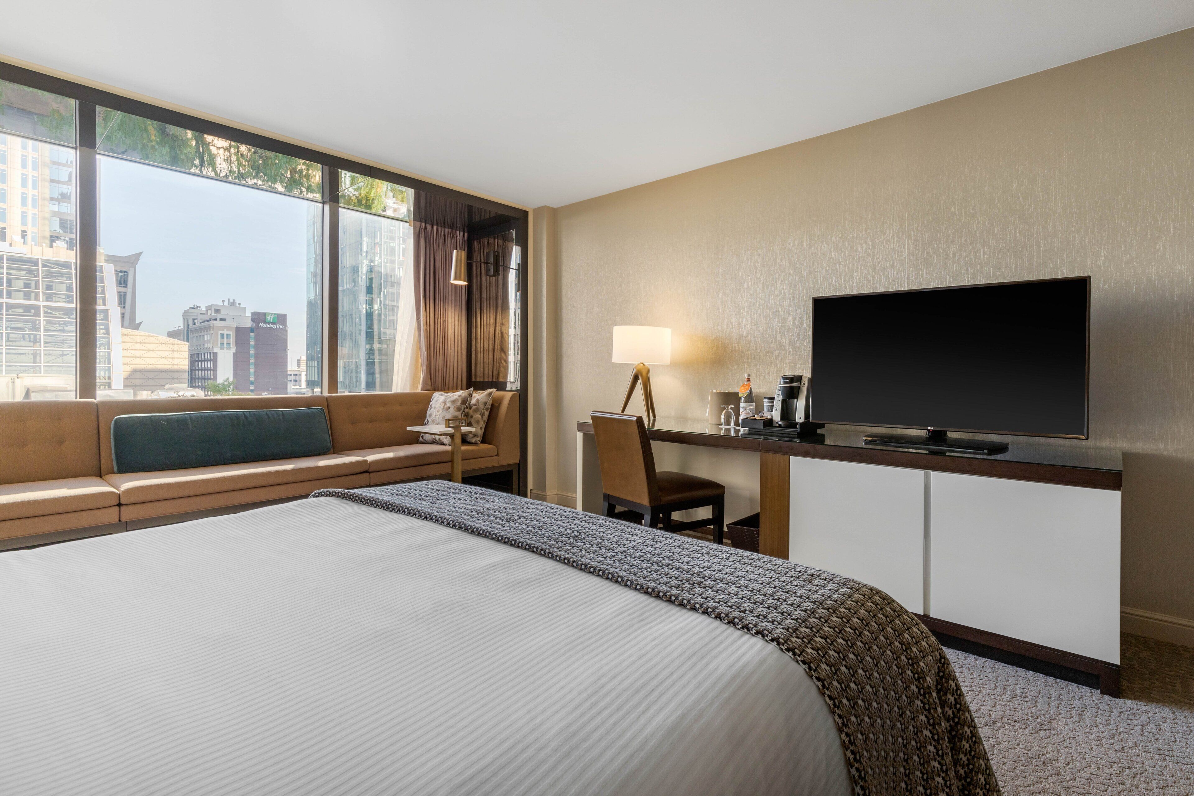 A hotel room with a large bed, a desk with a TV, a chair, a lamp, and a view of the city through large windows.