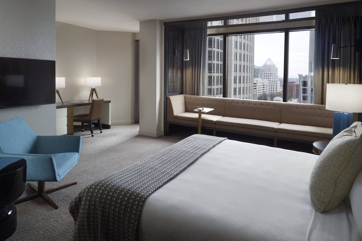 A hotel room with a large bed, a sofa, a desk with a chair, a TV, and a window with a city view.