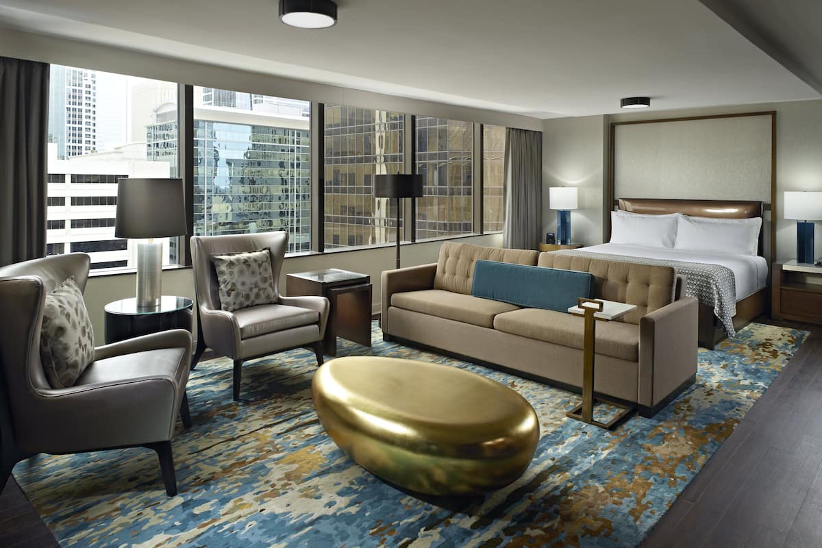 A hotel room with a large bed, a sofa, a chair, a small table, and a gold ottoman.