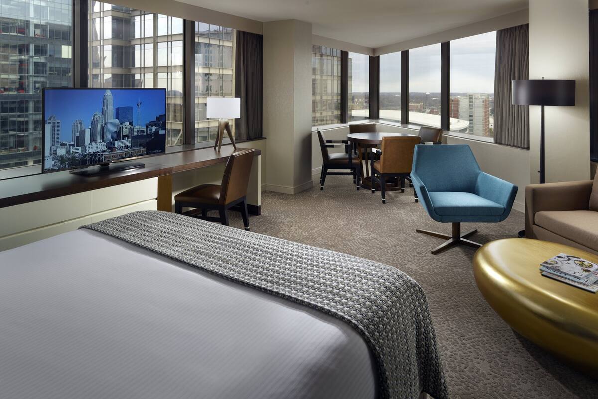A hotel room with a large bed, a desk with a TV, a dining area with chairs, and a view of the cityscape.