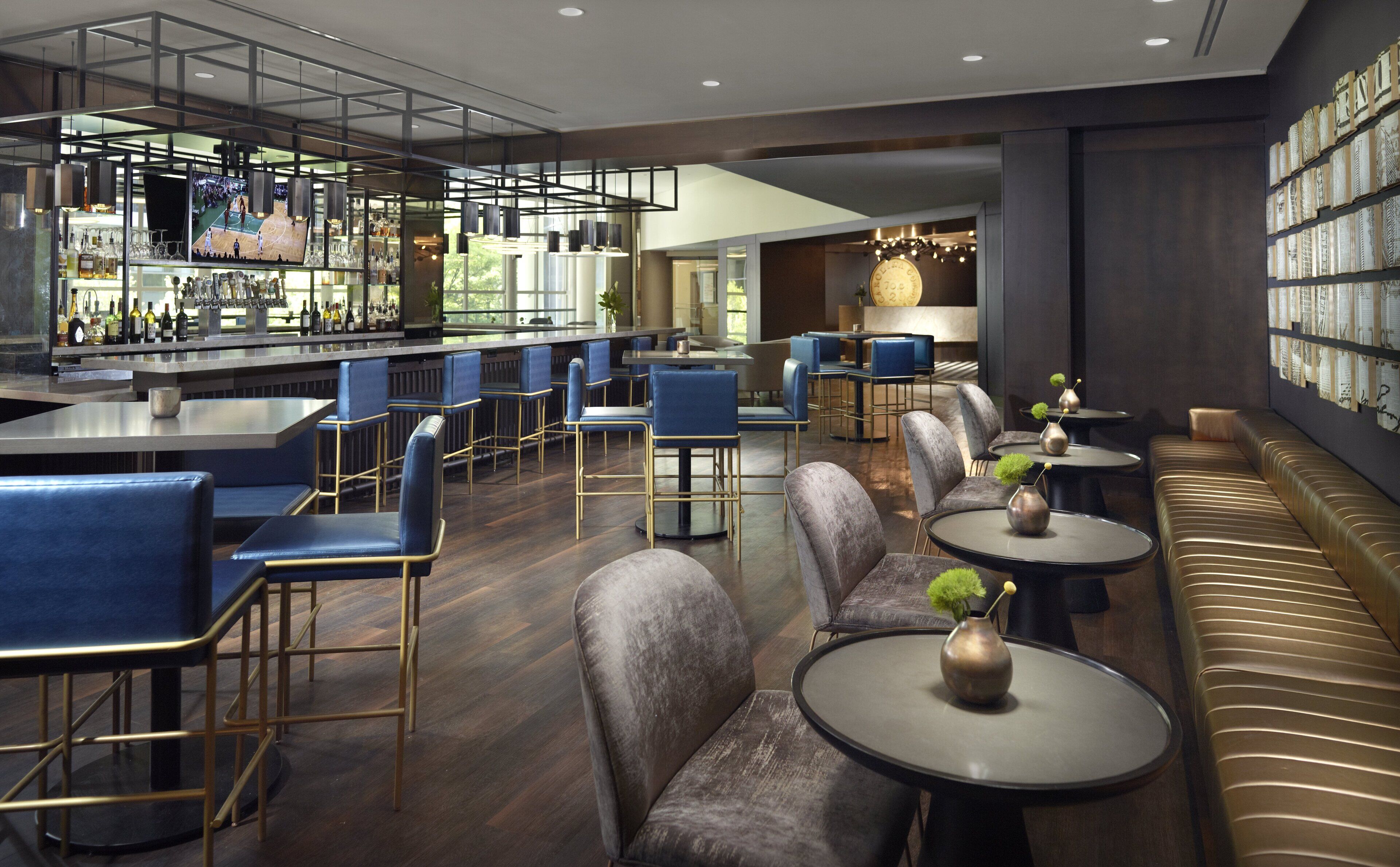 Un área de bar moderna con mesas altas y taburetes azules, un espacio de estar con mesas redondas y bancos acolchados, y una pared decorada con cuadros enmarcados.