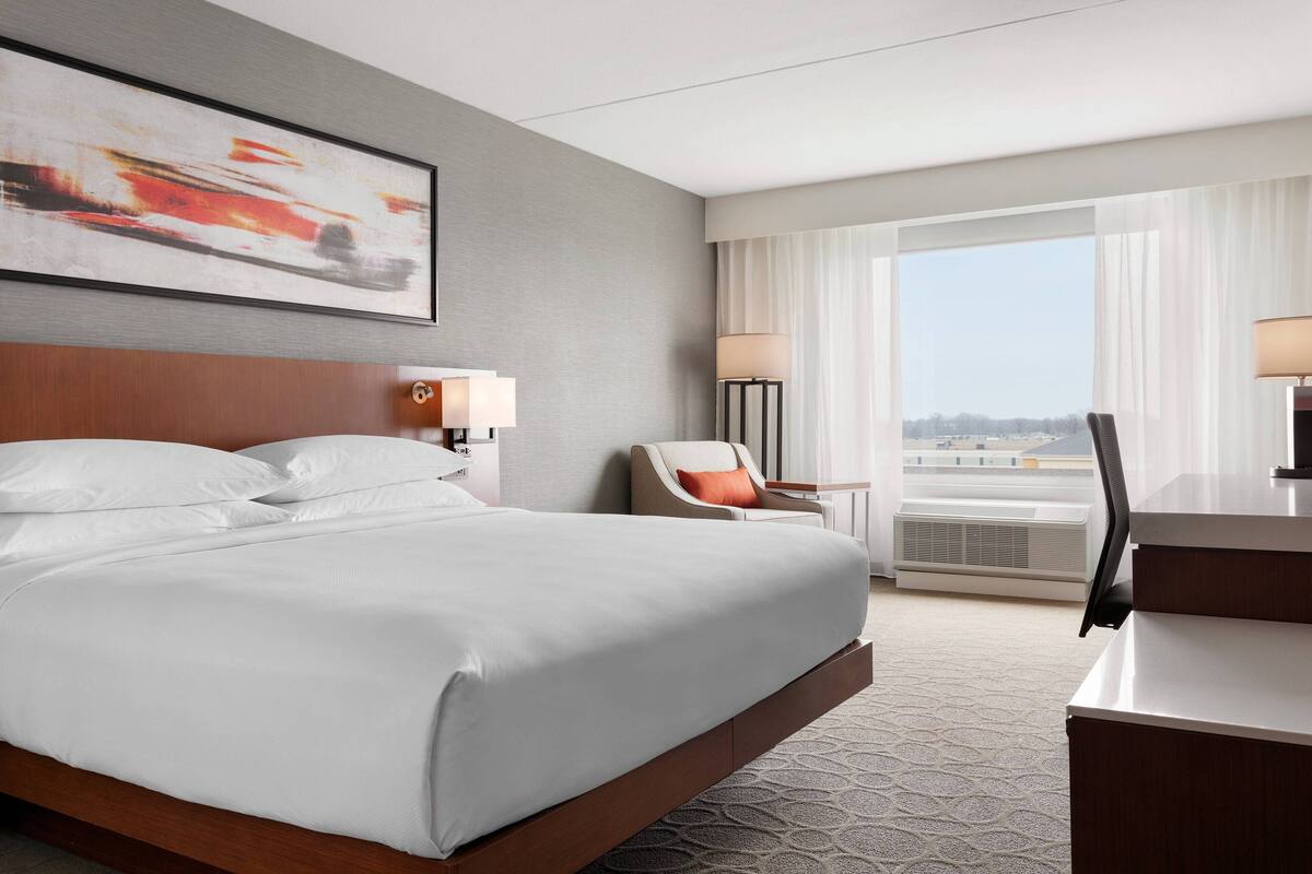 Une chambre d’hôtel avec un grand lit, un bureau, une chaise, une lampe et une vue sur la ville grâce à de grandes fenêtres.