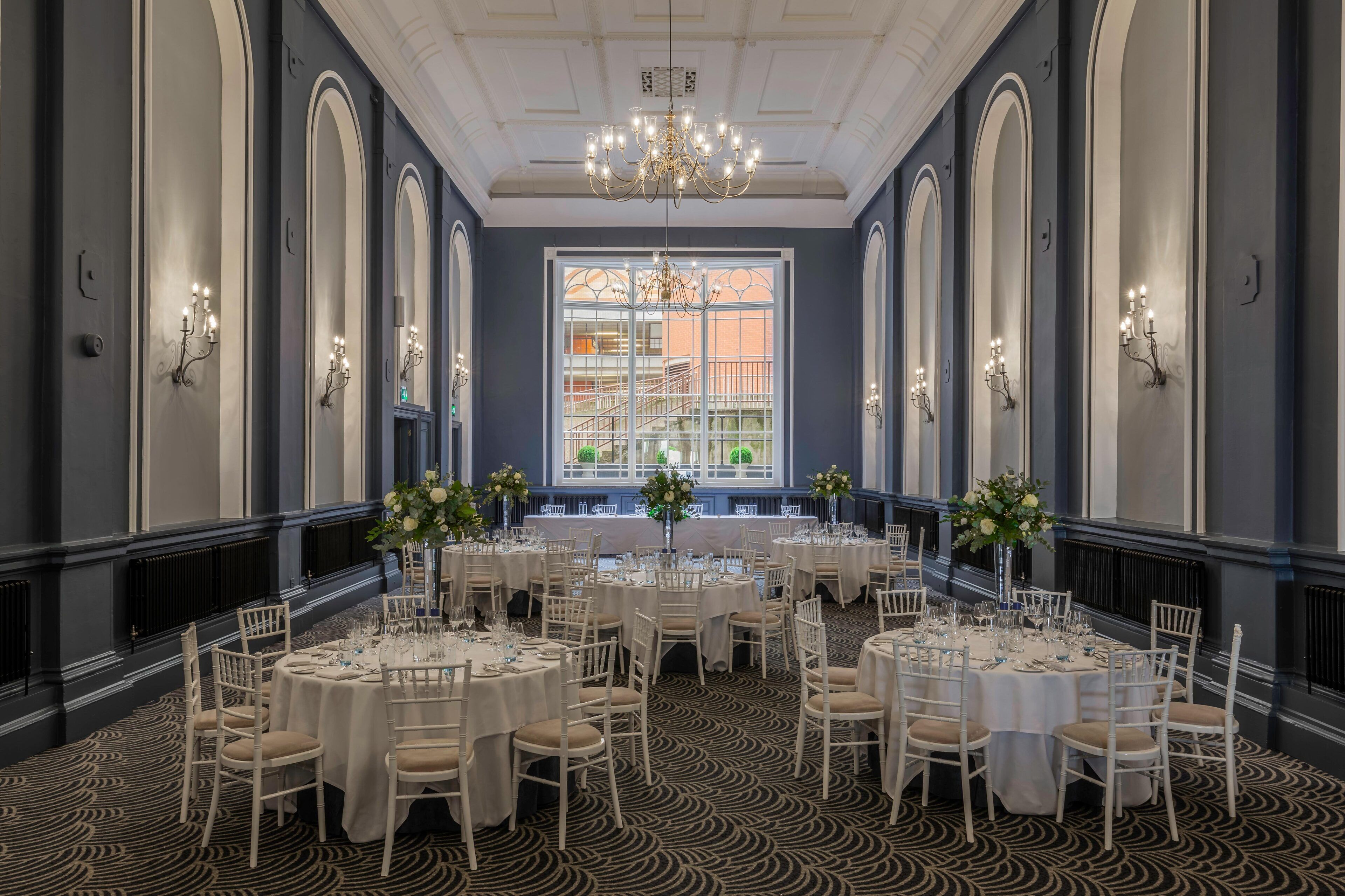 Une salle à manger élégante, avec plusieurs tables rondes dressées pour un repas, de grandes fenêtres offrant une vue dégagée et de magnifiques appliques murales.