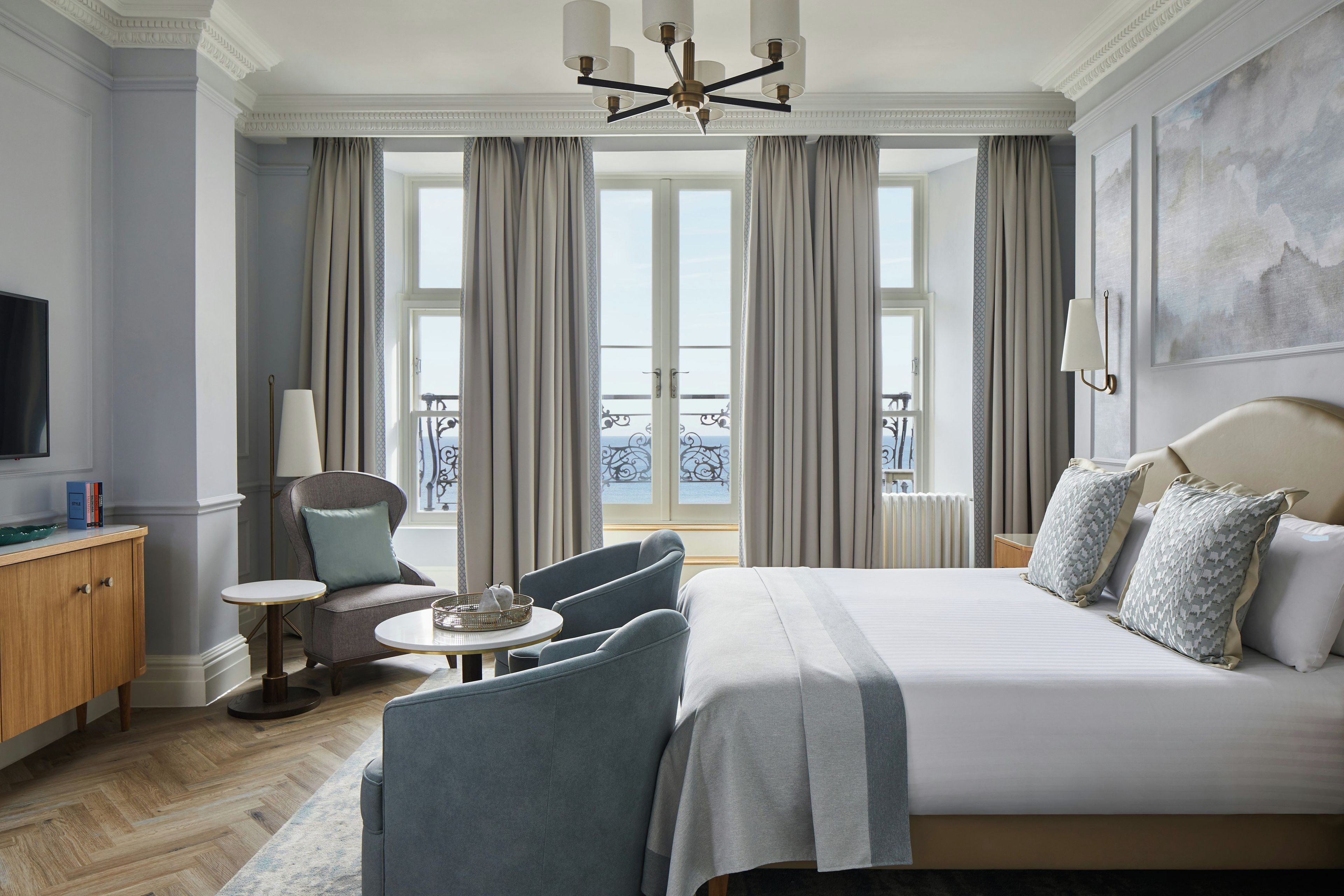 Habitación de hotel con una cama grande, televisor, zona de estar con una silla y una mesita, y vistas al exterior a través de las ventanas.