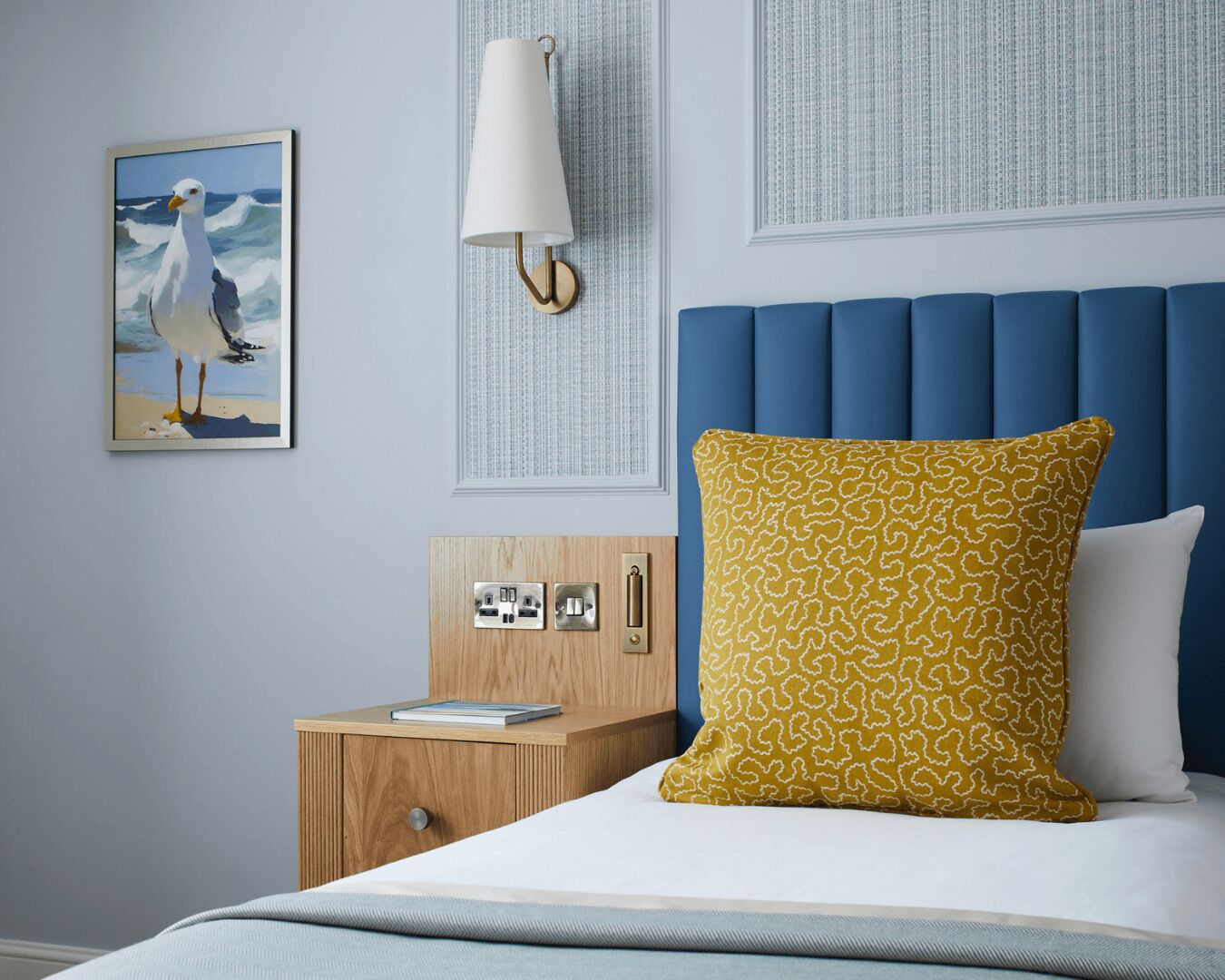 A bedroom with a blue headboard, a wooden nightstand with a lamp, a painting of a seagull, and a patterned pillow.