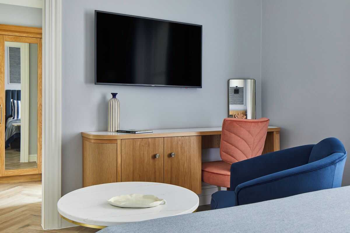 Une chambre d’hôtel moderne avec un téléviseur à écran plat fixé au mur, un bureau en bois agrémenté d’un vase, une chaise bleue et une table ronde avec une assiette.