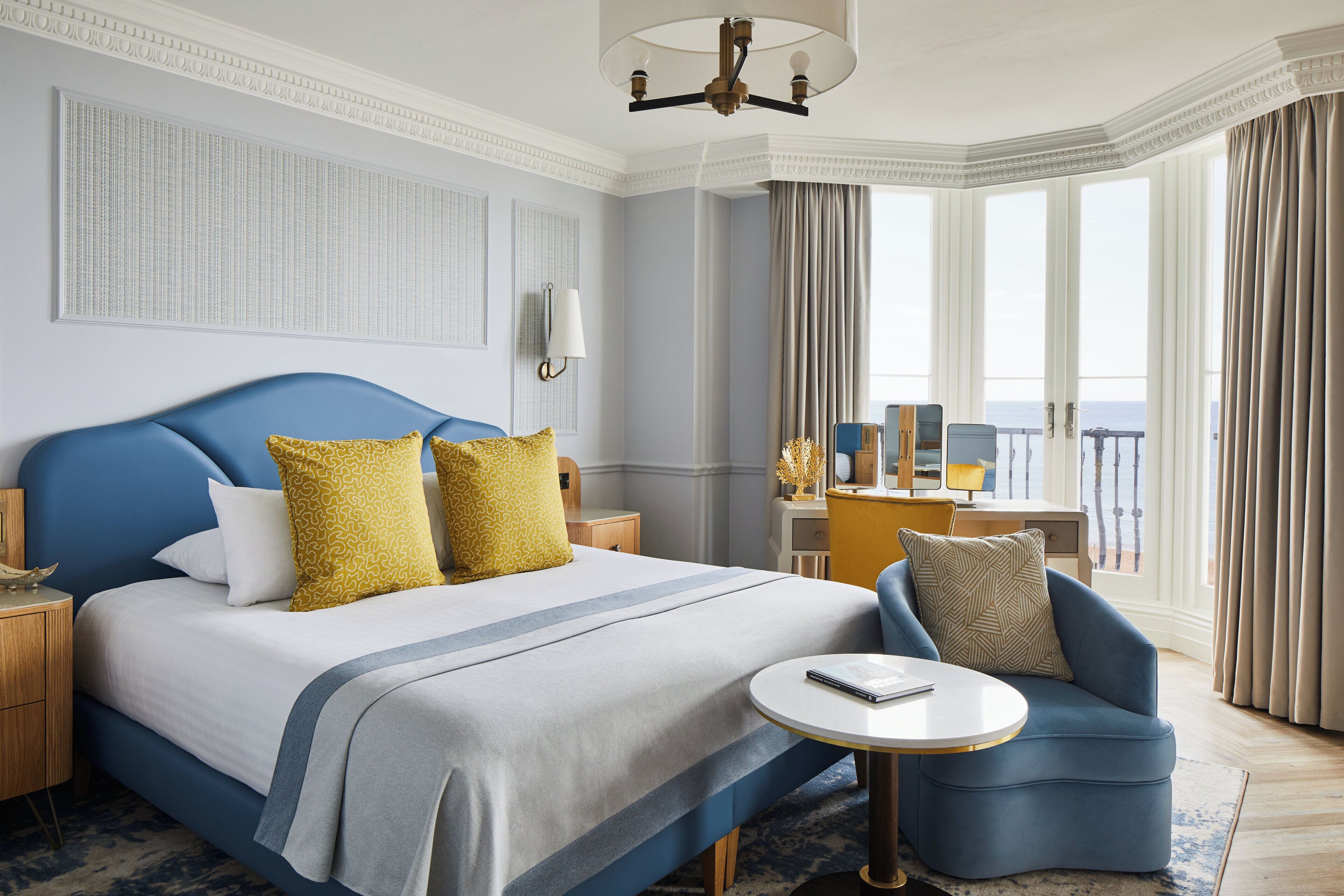 Habitación de hotel con una cama grande, un sillón azul, una mesa redonda y vistas al mar a través de amplios ventanales.