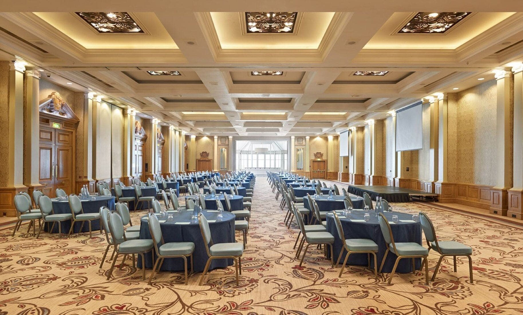 Un amplio salón de conferencias con filas de mesas y sillas, techos altos con detalles ornamentales y grandes ventanales que dejan entrar luz natural.