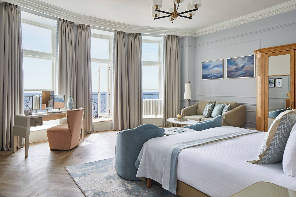 Une chambre d’hôtel spacieuse, dotée d’un grand lit, d’un bureau, d’une chaise et d’un canapé. De grandes fenêtres, équipées de rideaux, offrent une vue sur la mer, et une suspension en cristal illumine la pièce.