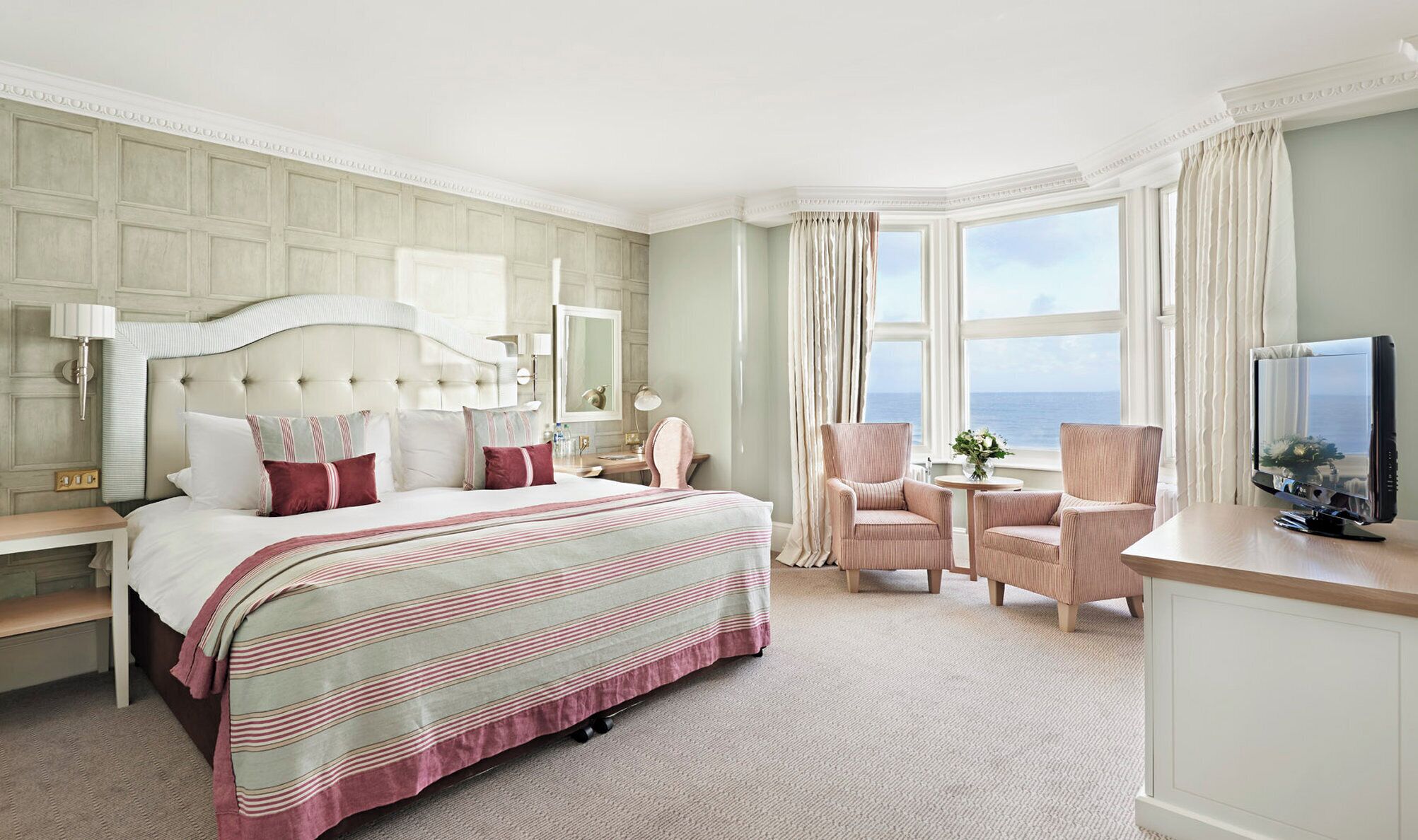 A hotel room with a large bed, a desk with a TV, two chairs, a small table, and a view of the sea.