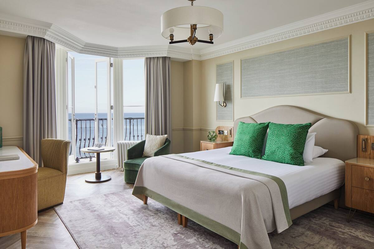 A hotel room with a large bed, a desk, a chair, and a view of the sea through the windows.