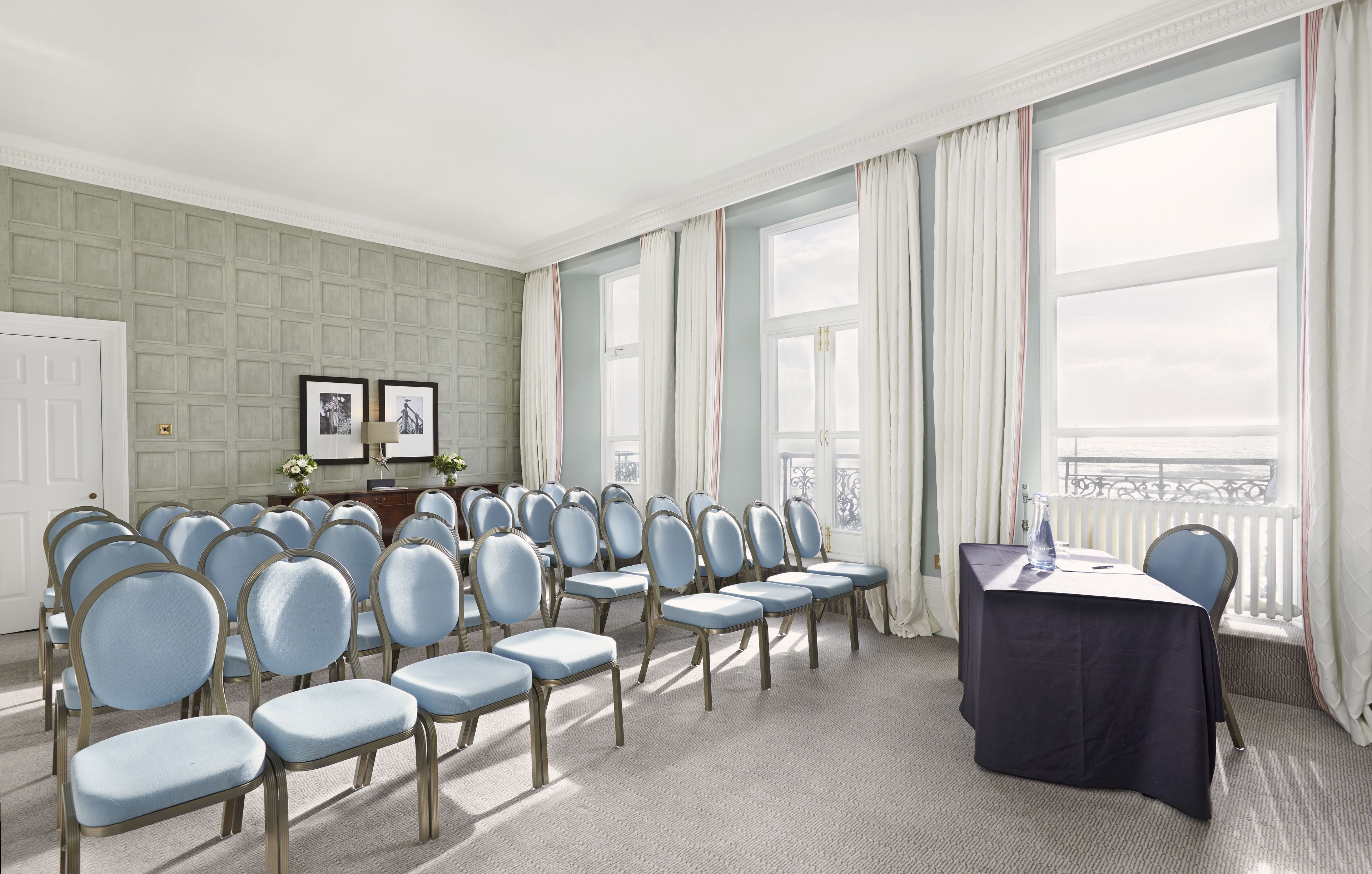A conference room with rows of chairs, a table with a water bottle, and large windows with curtains.