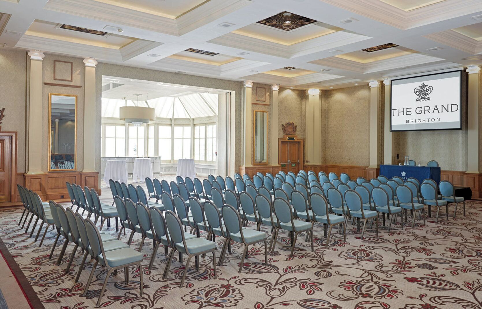 A conference room with rows of chairs, a large screen, and a banner reading 'THE GRAND BRIGHTON'.