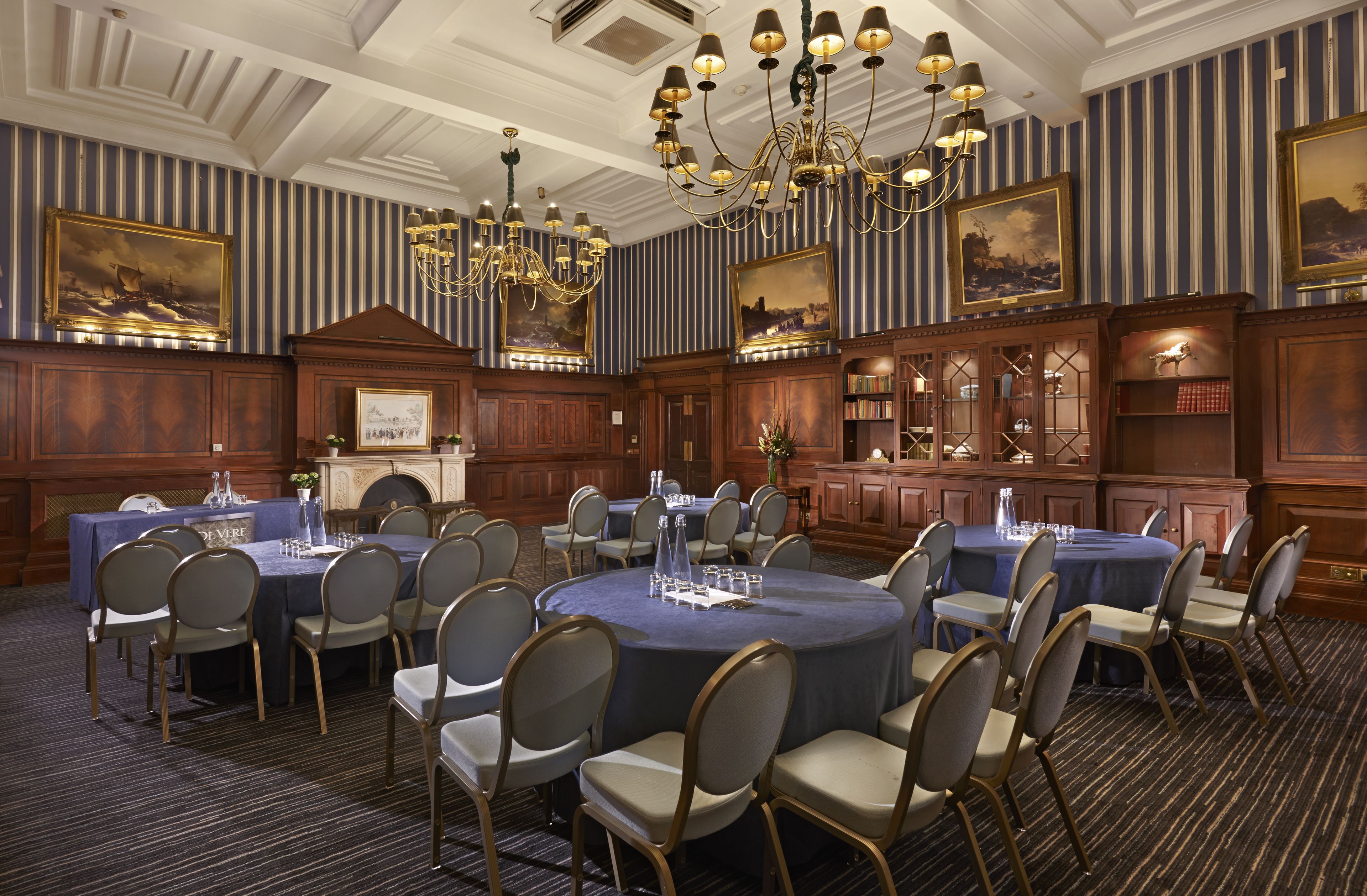 A formal dining room with round tables, chairs, chandeliers, and framed artwork on the walls.