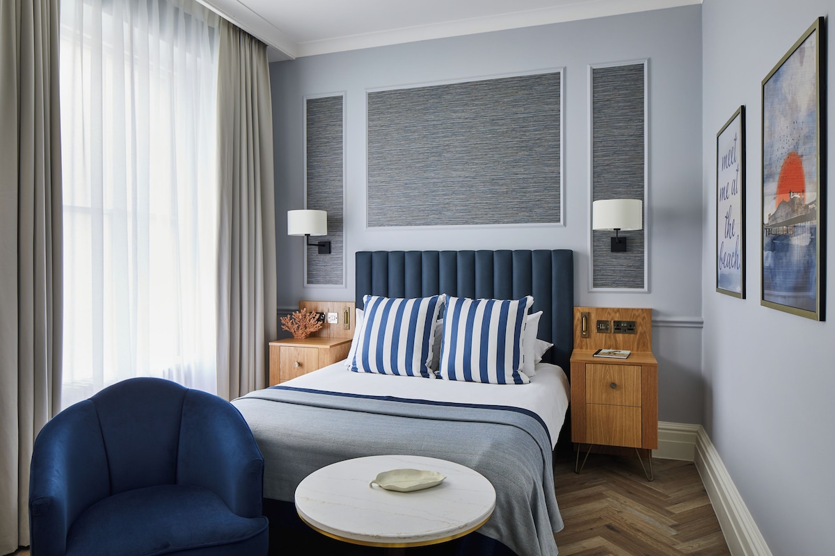 A hotel room with a bed, a round table, a chair, a nightstand, and a lamp.