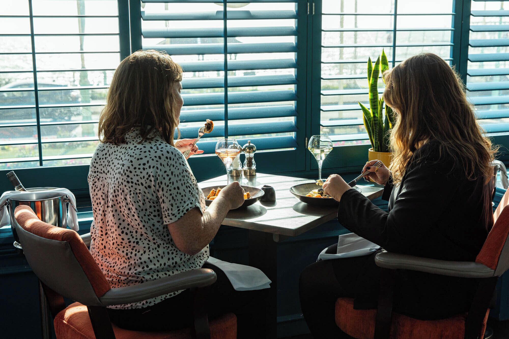 Deux personnes sont assises à une table, près d’une grande fenêtre donnant sur l’extérieur, et y prennent leur repas.