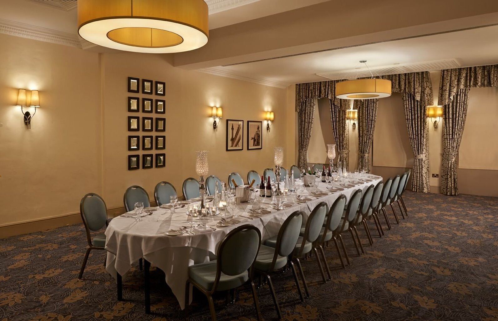 A conference room with a long table set for a formal event, surrounded by chairs, with wall-mounted lighting and framed pictures.