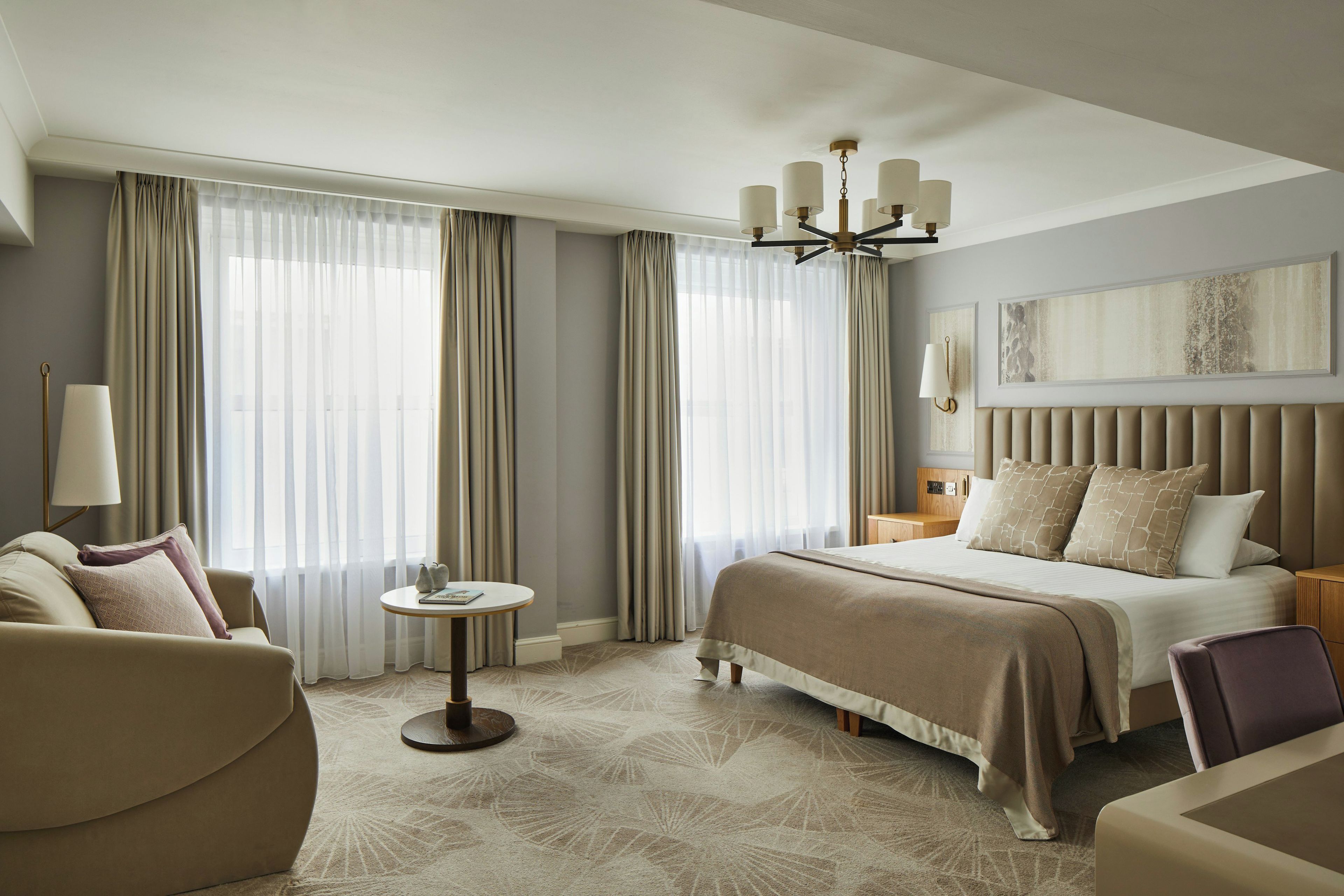 A hotel room with a large bed, a sitting area with a chair and a small table, and a large window with curtains.