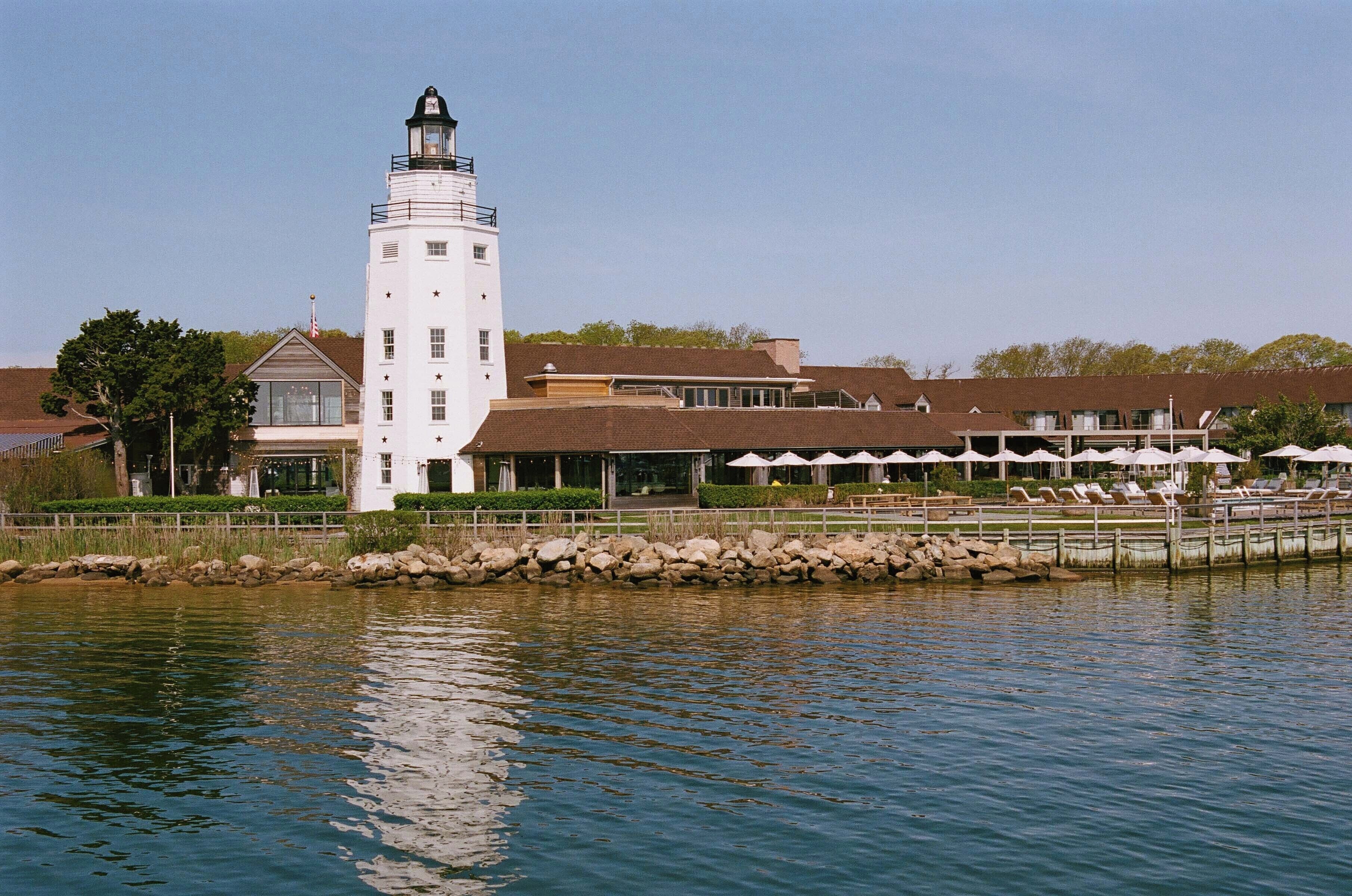 A white lighthouse with a tower and a base, a building with a brown roof, and a waterfront area with lounge chairs and umbrellas.