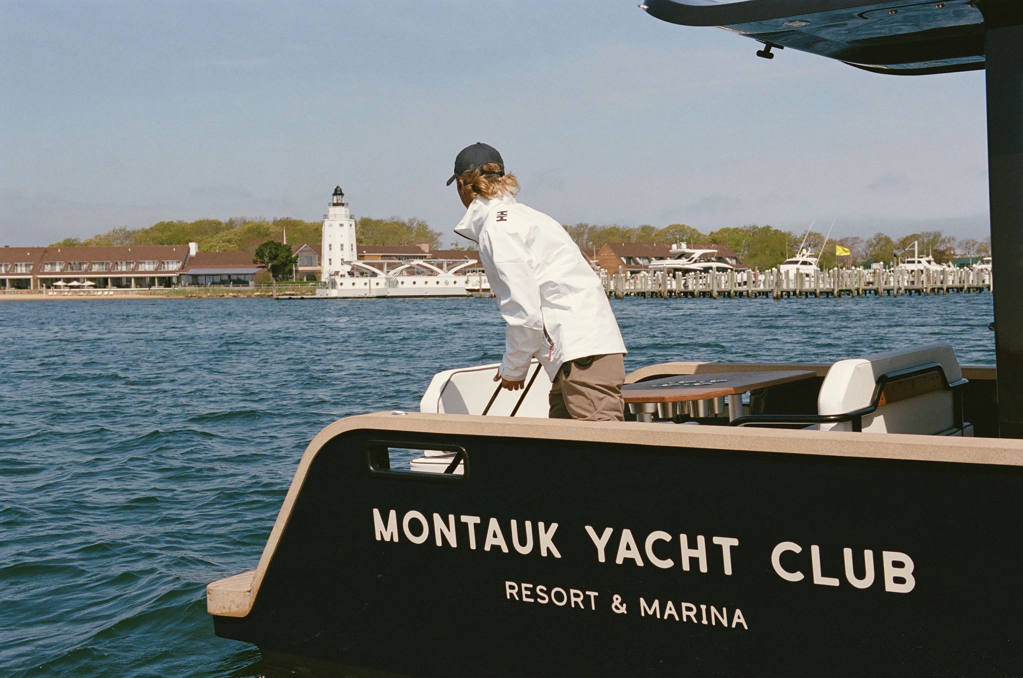 A boat with "Montaouk Yacht Club" written on the side, docked near a lighthouse and marina with other boats.