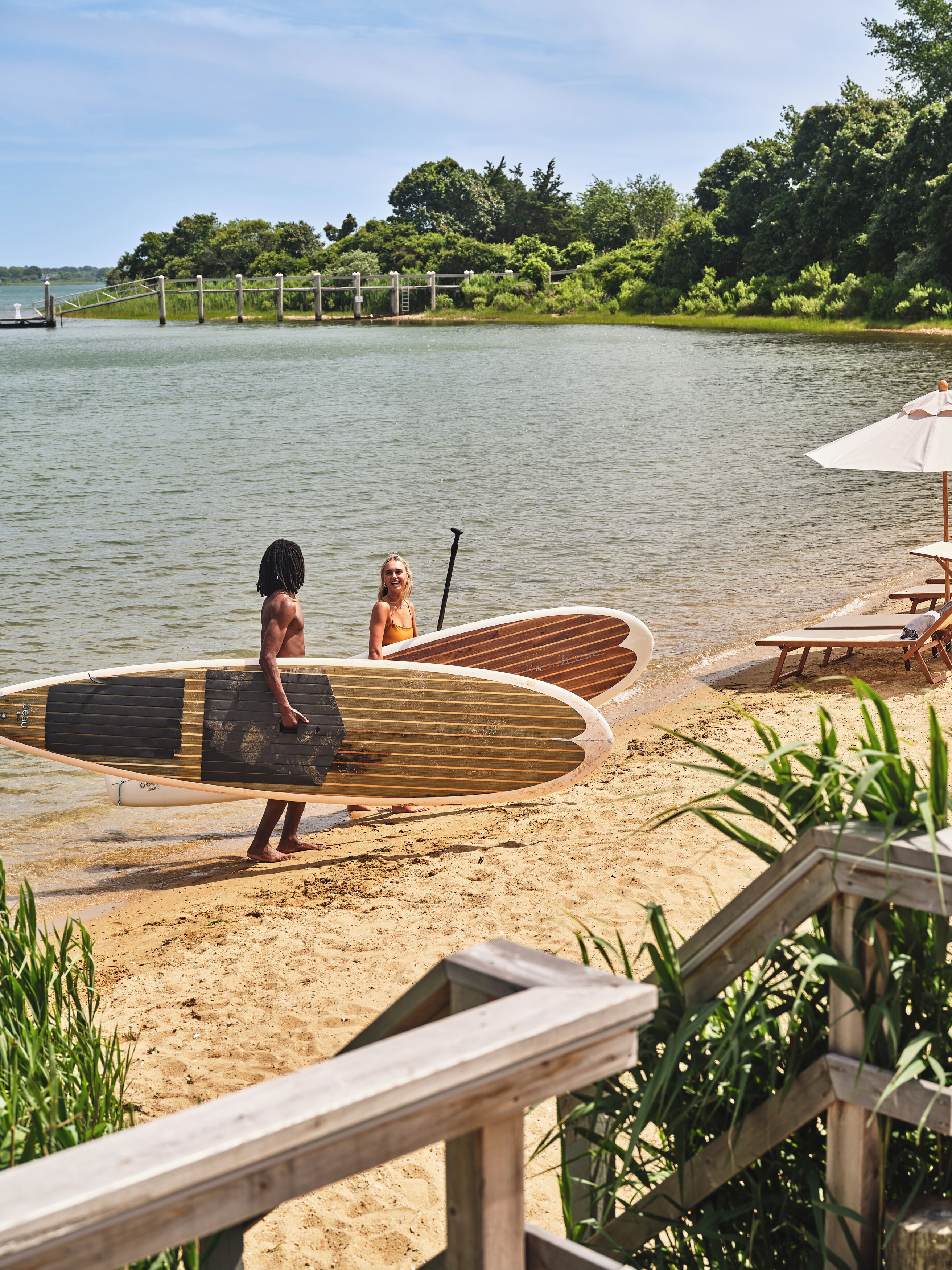 A wooden deck leading to a sandy beach with a calm lake in the background.