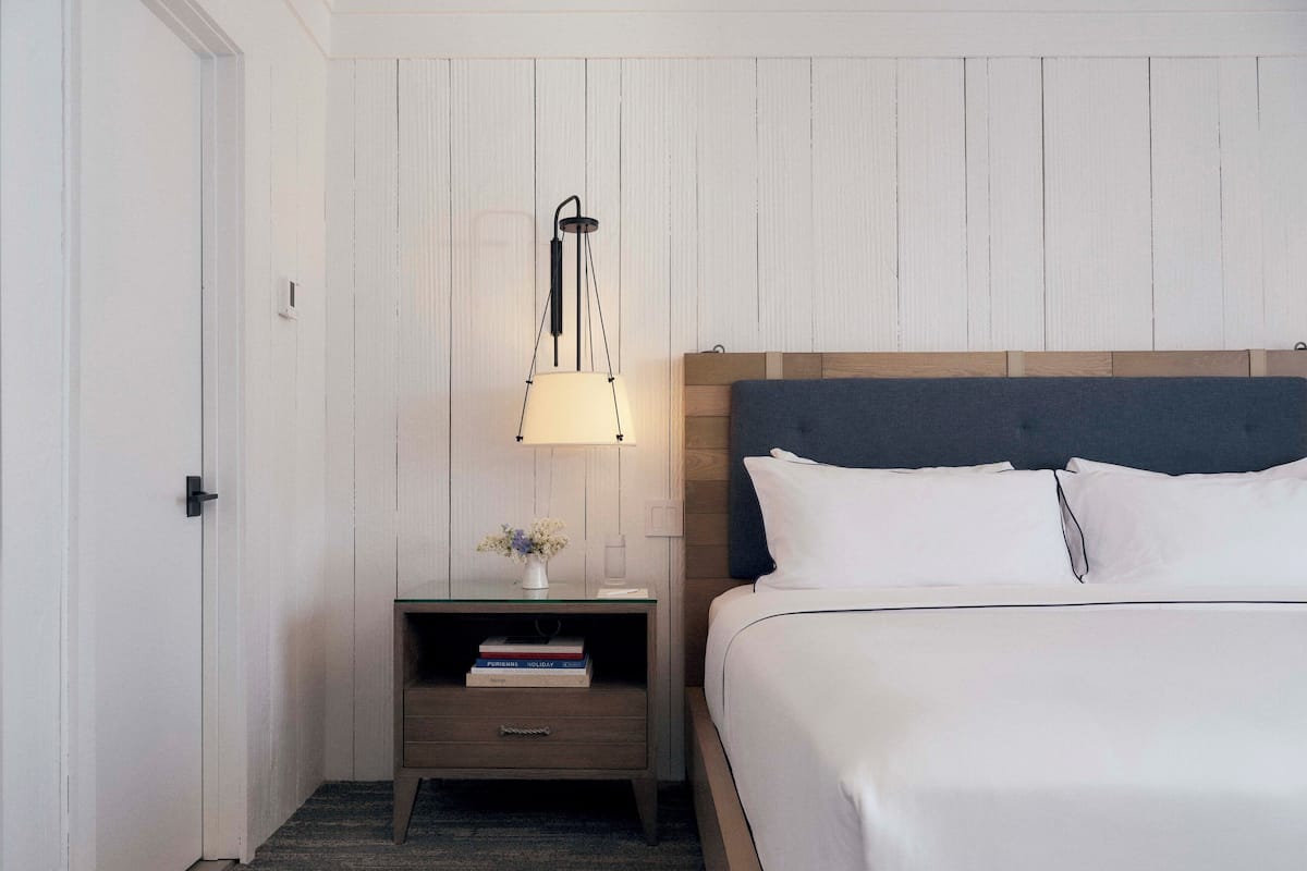 A hotel room with a bed, a nightstand with a lamp, and a small table with a vase of flowers and books.