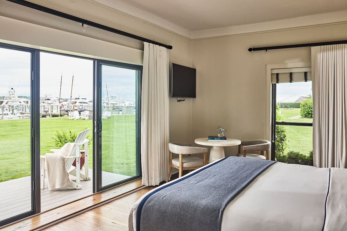 A hotel room with a large sliding glass door leading to a balcony, a bed with a blue blanket, a small round table with a chair, and a TV mounted on the wall.