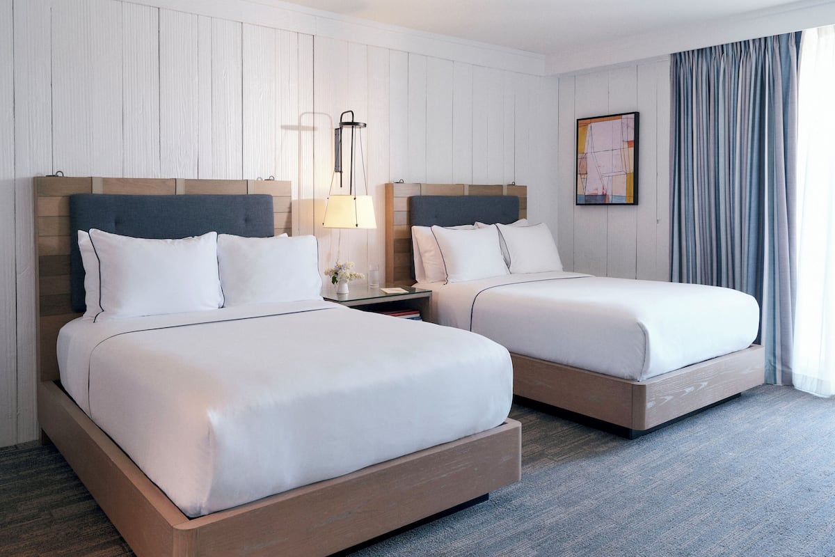 A hotel room with two beds, a nightstand, a lamp, a framed picture on the wall, and a window with curtains.