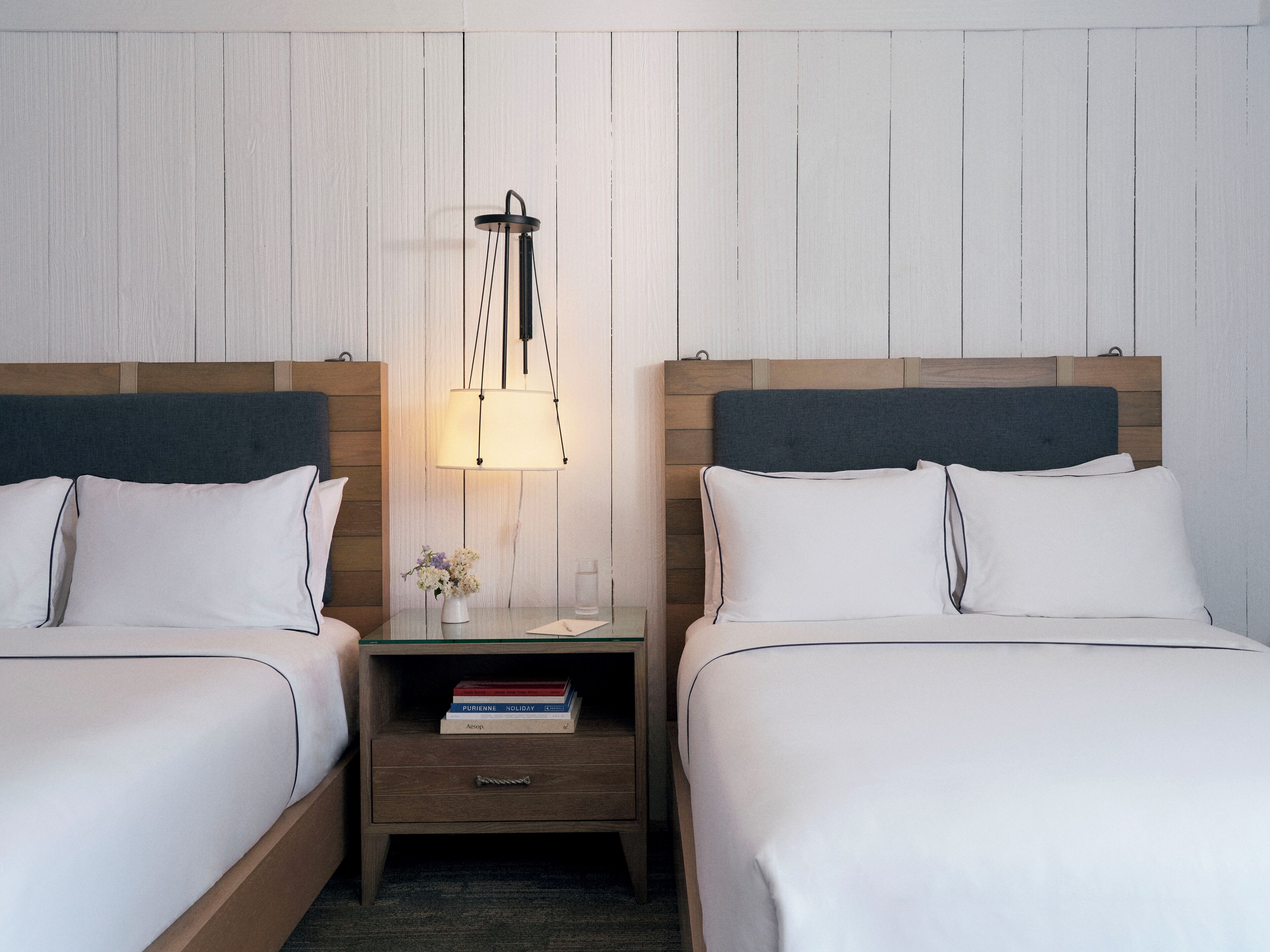 A hotel room with two single beds, a bedside table with a lamp, a small nightstand, and a vase with flowers.
