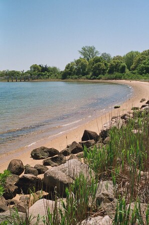 Une plage à l’eau claire, avec un rivage sablonneux et de gros rochers.
