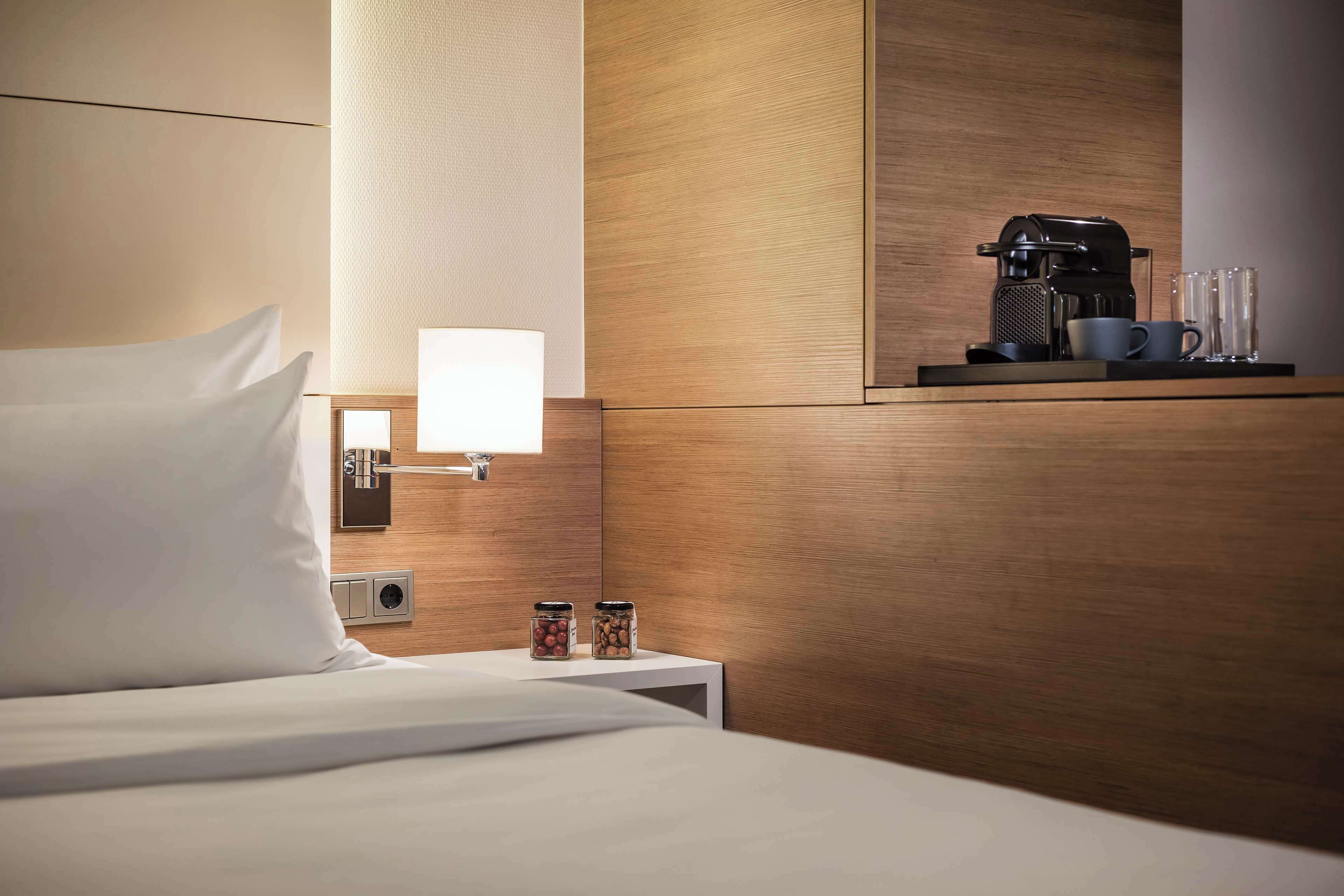 Habitación de hotel con una cama bien hecha, una mesita de noche con lámpara, una cafetera y dos recipientes con aperitivos.