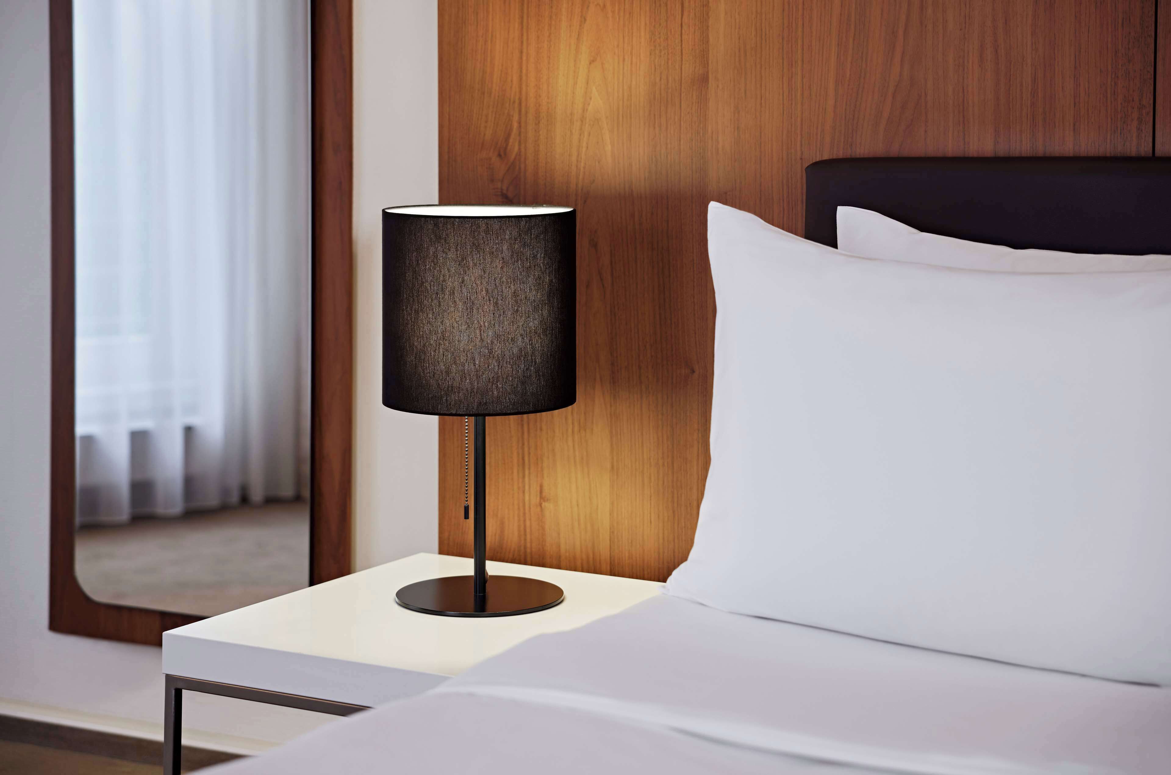 A neatly made bed with white linens and a black headboard, a modern table lamp with a dark shade, and a wooden panel wall in the background.