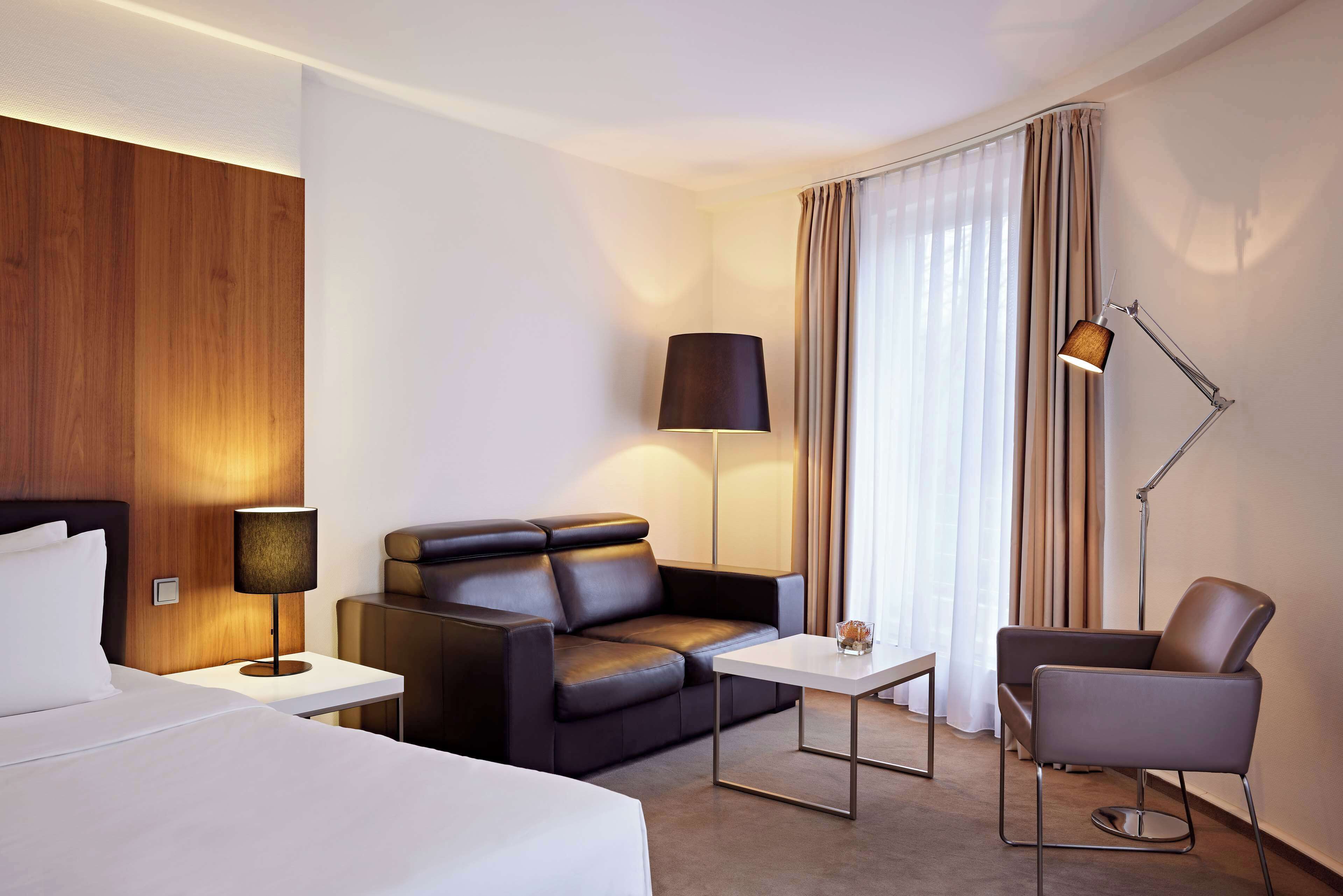 Una habitación de hotel moderna con cama, sofá de cuero, una mesita, una silla y una lámpara de escritorio.
