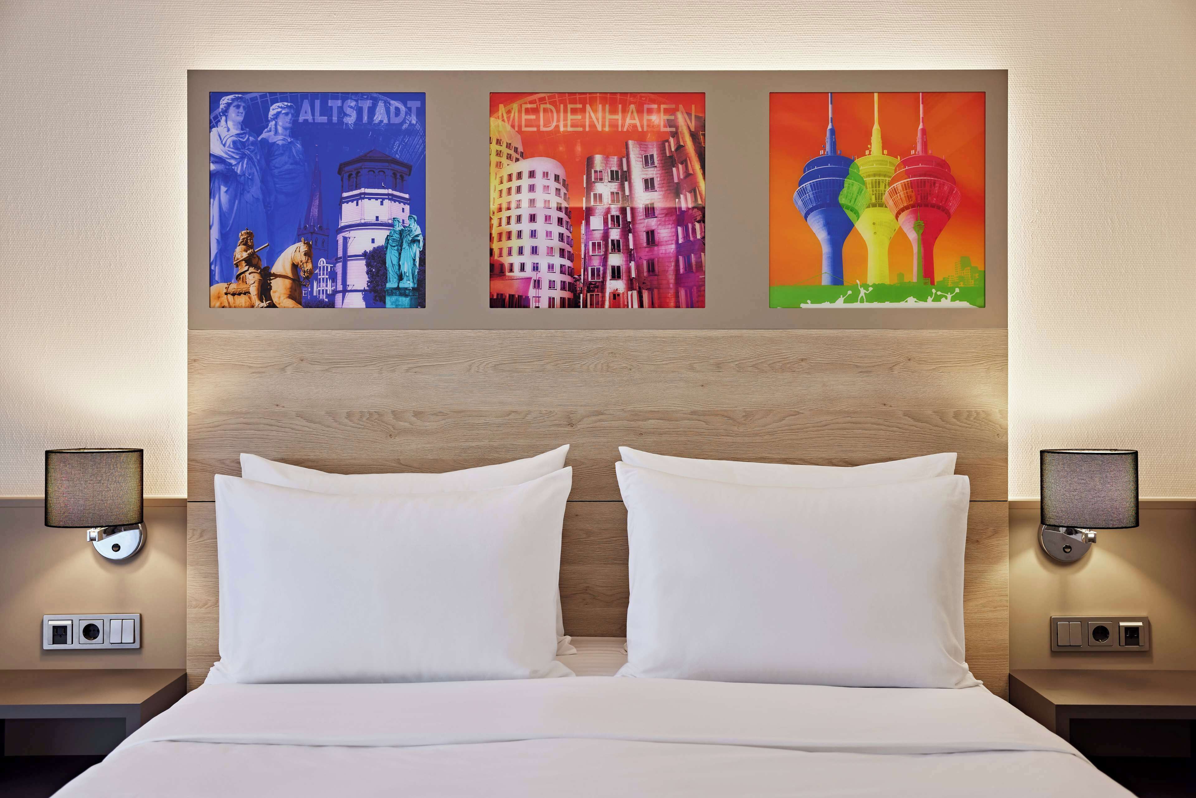 Quarto de hotel com uma cama, duas mesas de cabeceira e uma parede com três pinturas coloridas de paisagens urbanas.