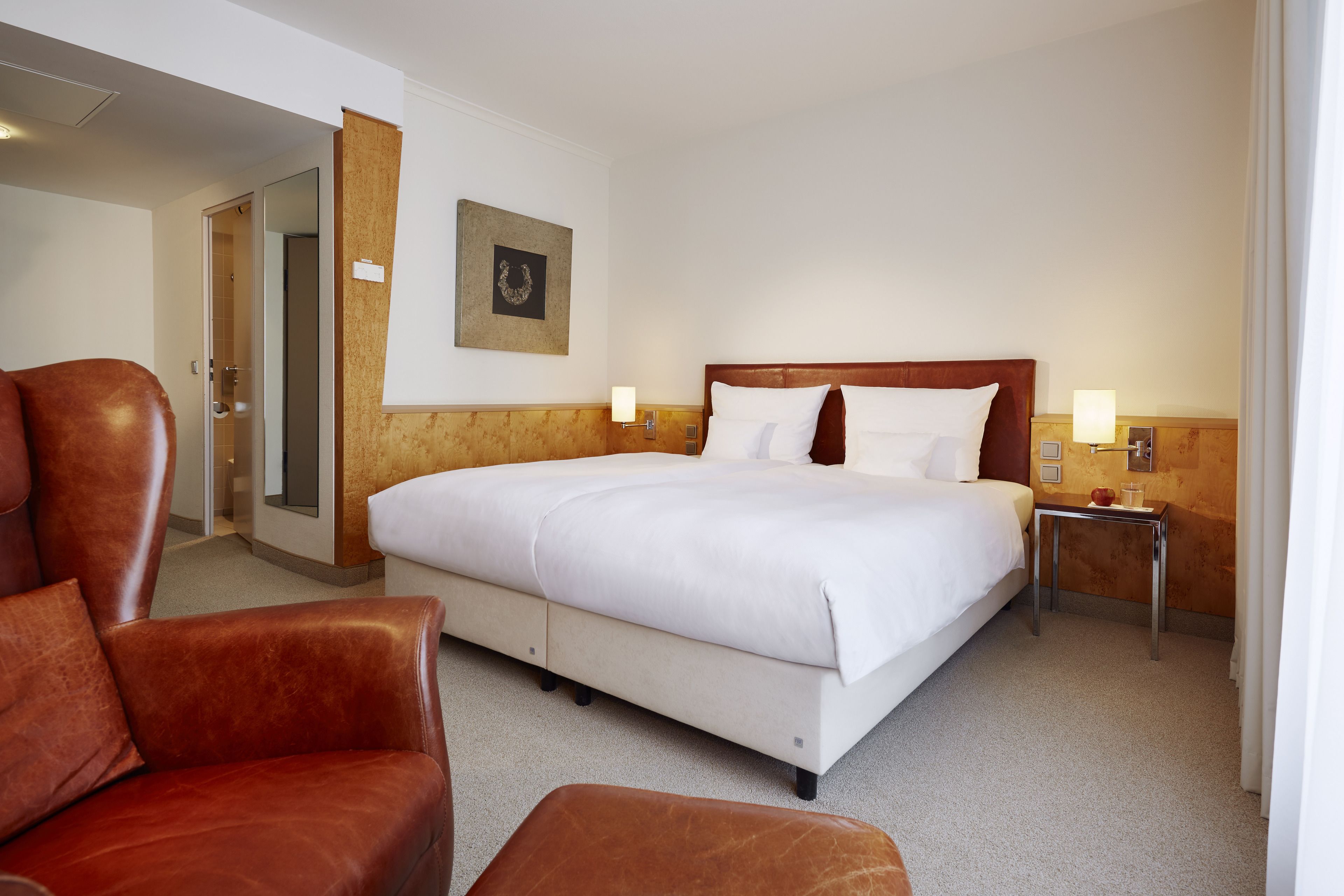 Una habitación de hotel con una cama grande, un cabecero de madera, dos mesitas de noche con lámparas, un sillón de cuero marrón y una puerta que da a un baño.