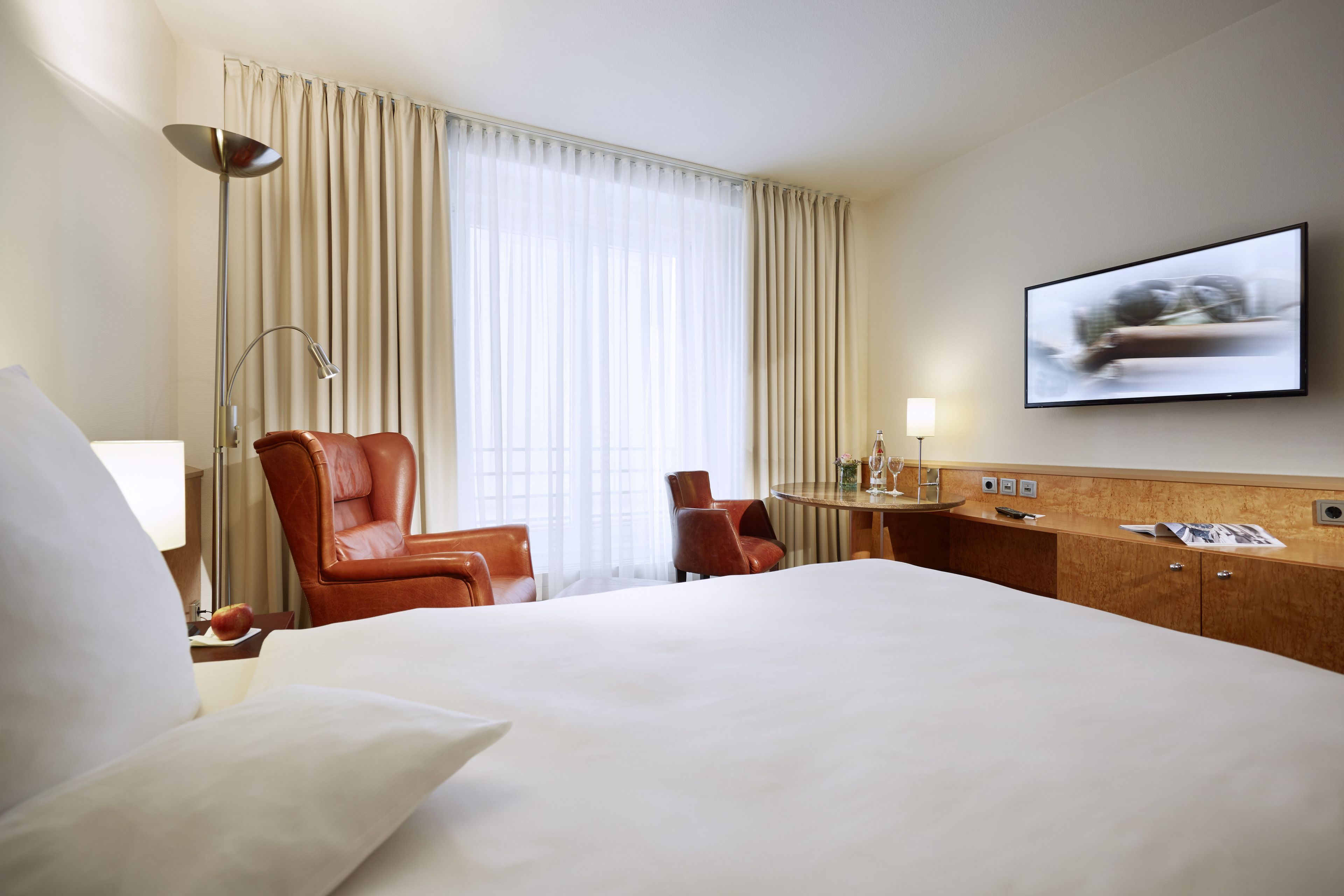 A hotel room with a large bed, two leather chairs, a desk with a lamp, a television, and a window with curtains.