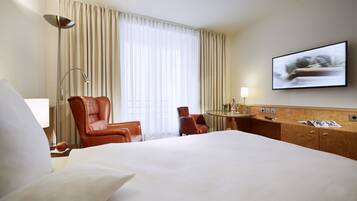 A hotel room with a large bed, two leather chairs, a desk with a lamp, a television, and a window with curtains.