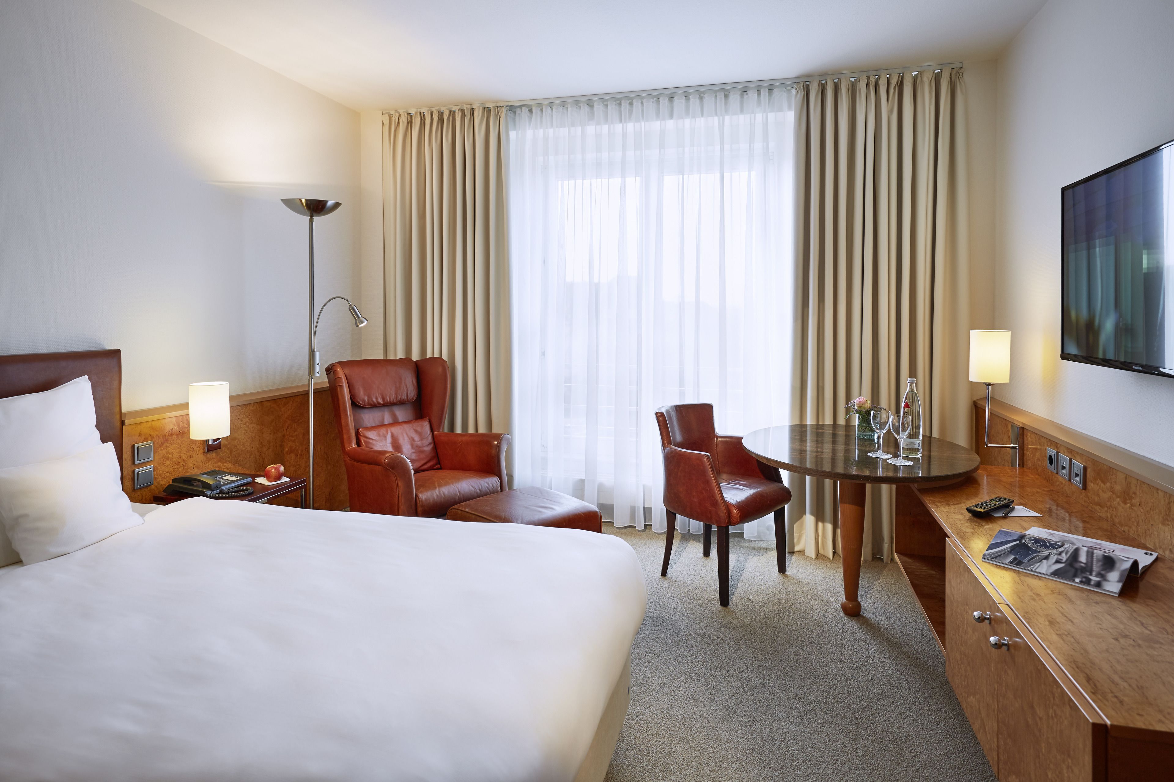 Ein Hotelzimmer mit einem großen Bett, einem Ledersessel, einem kleinen Tisch mit Lampe und einem an der Wand befestigten Fernseher.