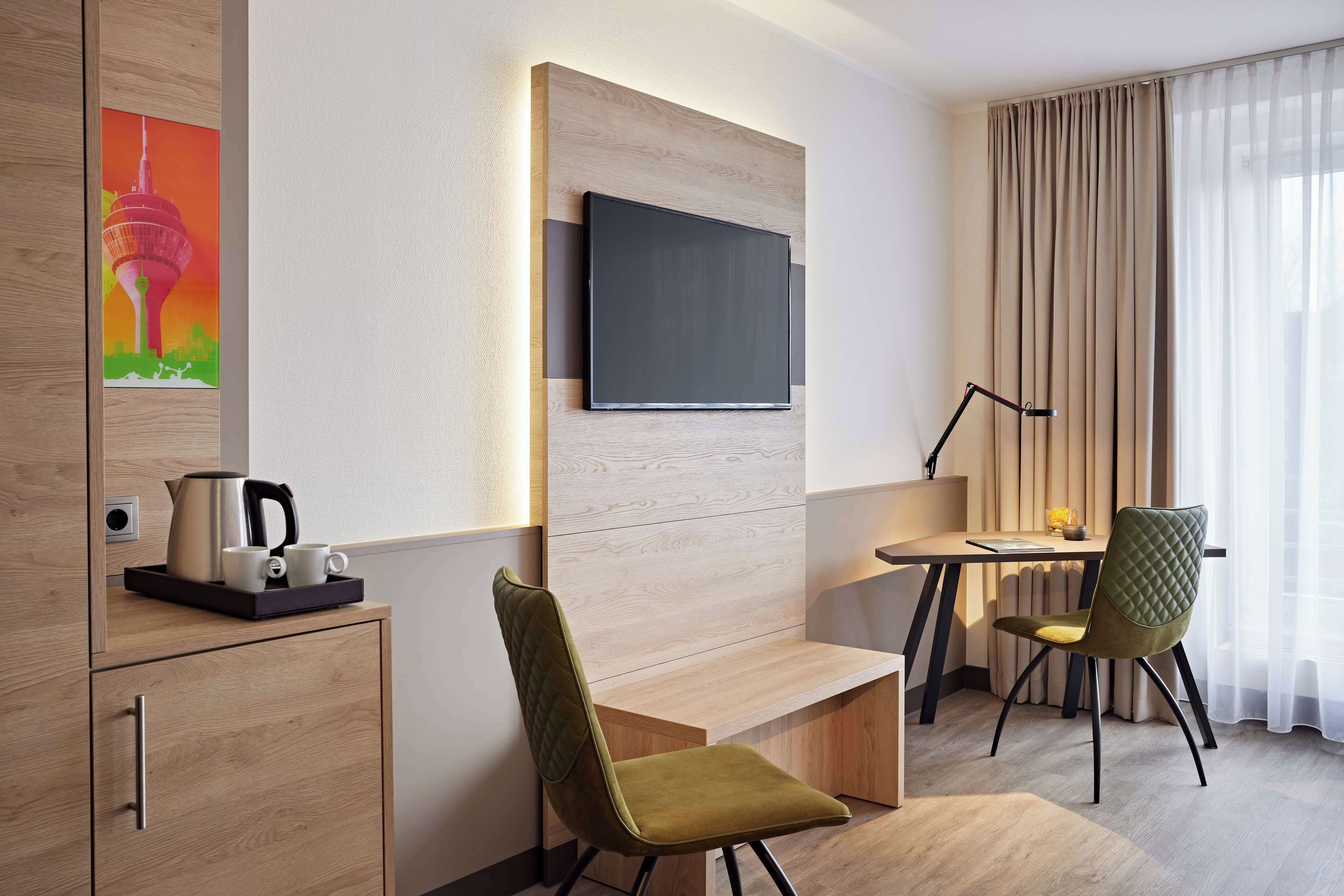 Habitació d'hotel moderna amb televisió de pantalla plana penjada a la paret, escriptori de fusta amb una làmpada, cadira tapissada de color verd i una petita zona de cuina.