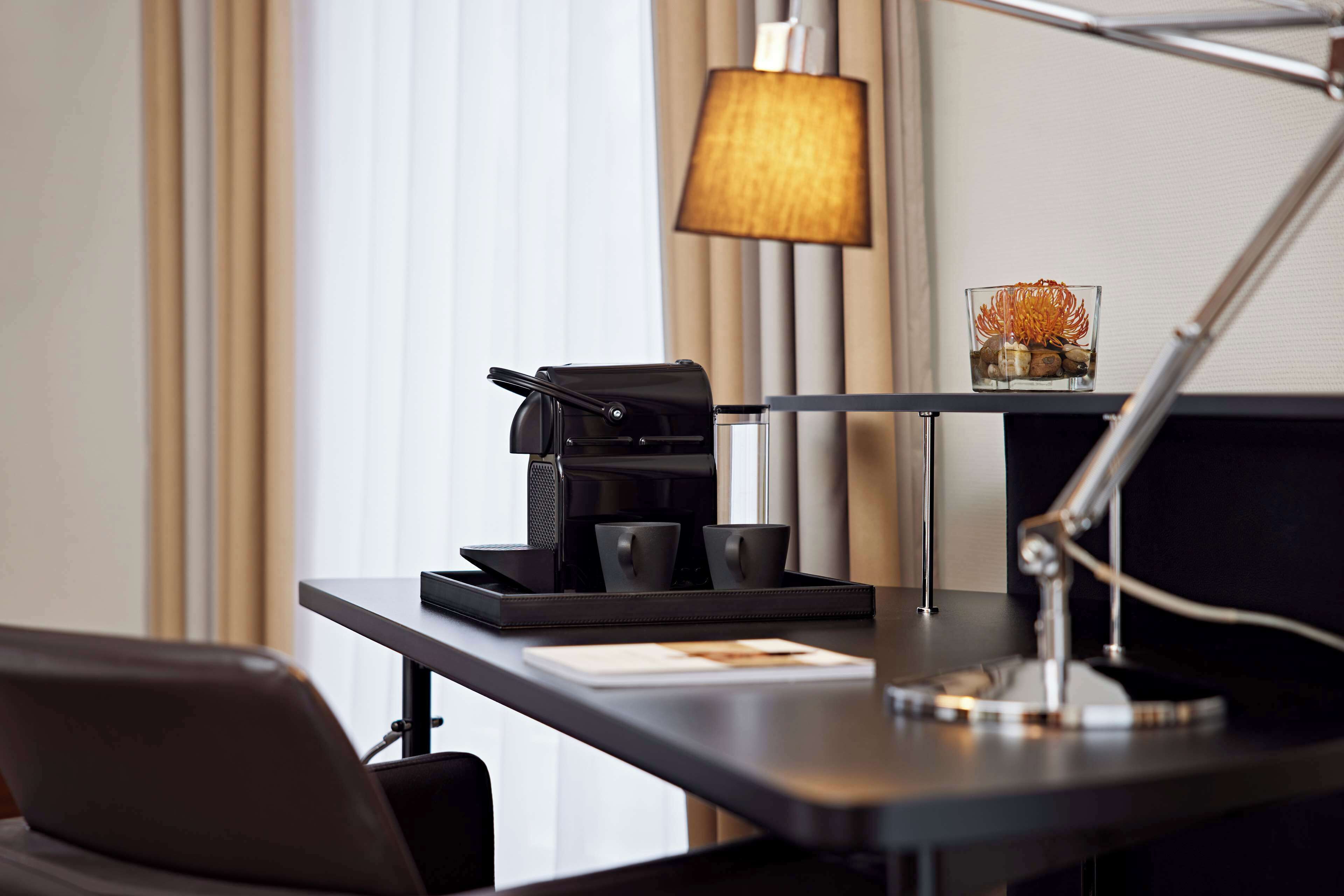 A hotel room desk with a coffee maker, two cups, a lamp, and a decorative glass.