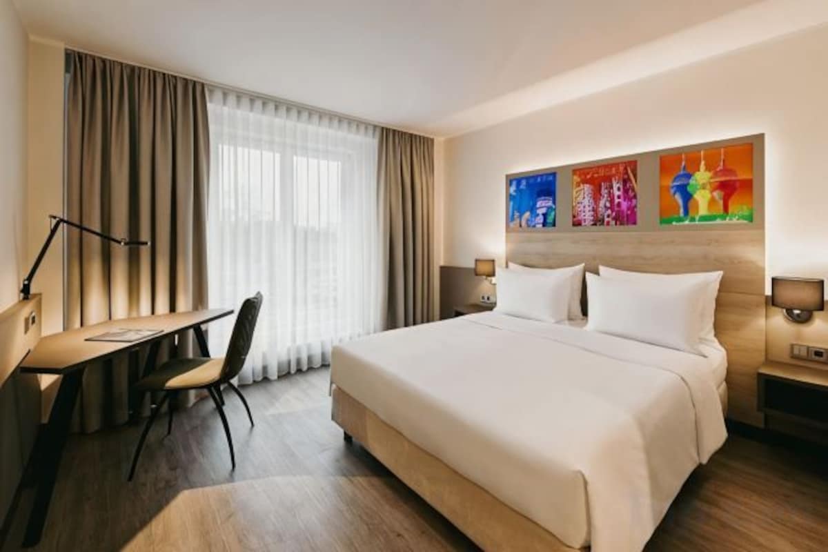 A hotel room with a bed, desk, chair, and artwork on the wall.