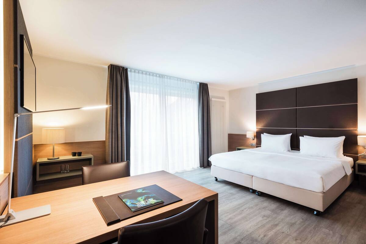 Une chambre d’hôtel avec un lit, un bureau avec un ordinateur portable, une chaise, une table de chevet, une lampe et une fenêtre avec des rideaux.