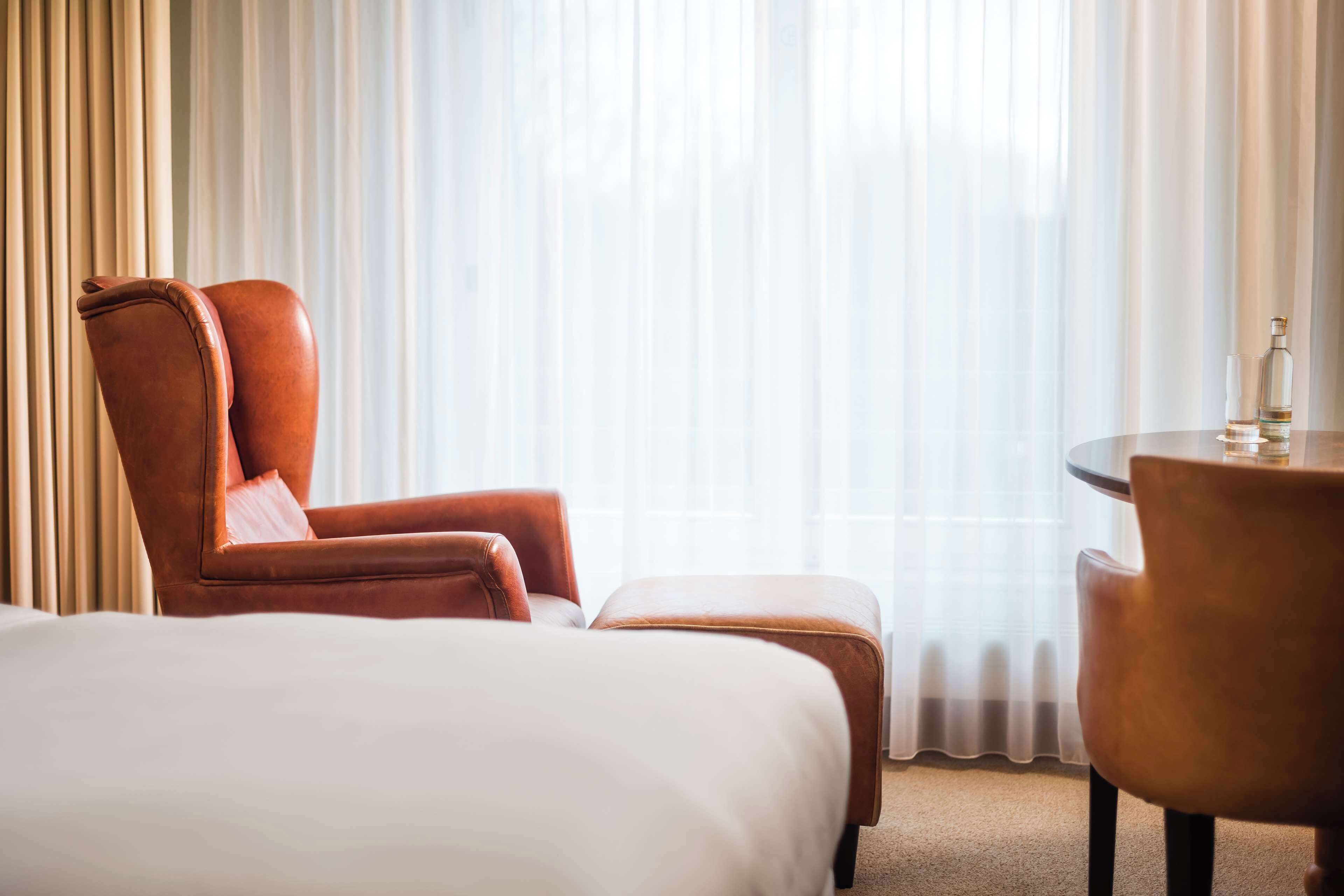 Una habitación de hotel con una cama grande, un sillón marrón, una mesita con una botella y cortinas traslúcidas.