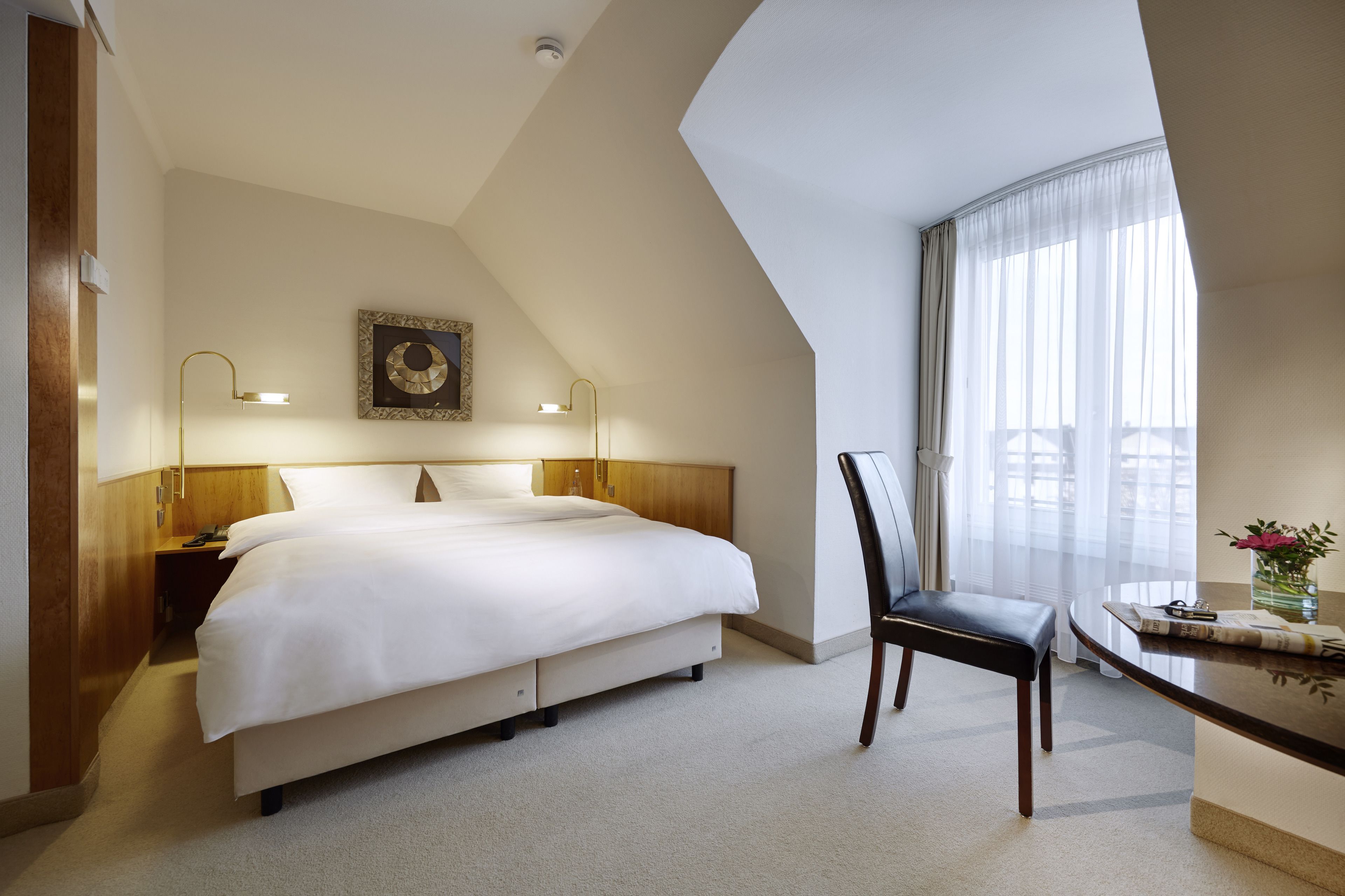 Ein Hotelzimmer mit einem großen Bett, einem Schreibtisch mit Stuhl, einem Fenster mit Vorhängen und einer Wandlampe.