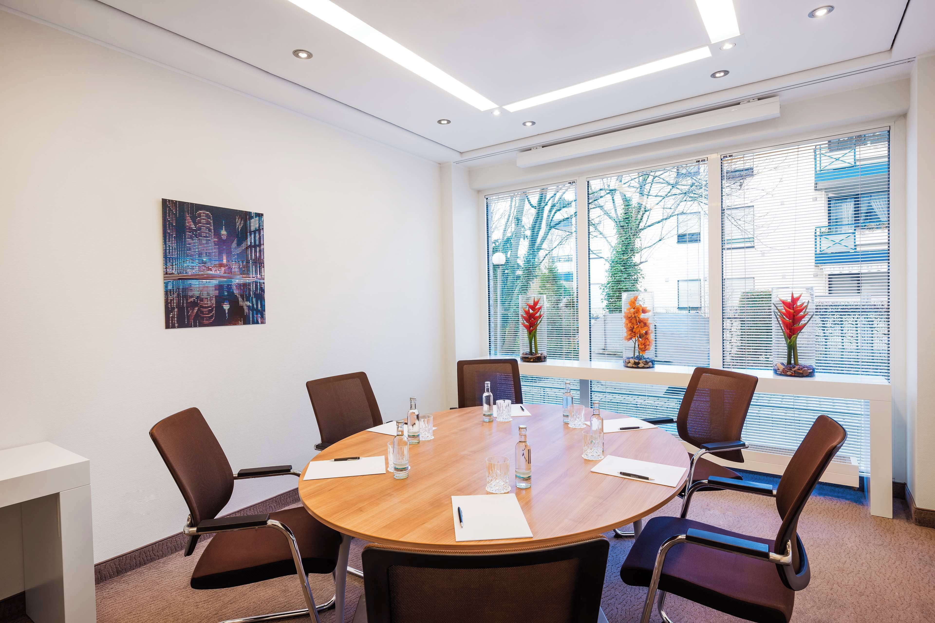Une salle de conférence équipée d’une table ronde, de six chaises et d’une grande fenêtre avec des stores.