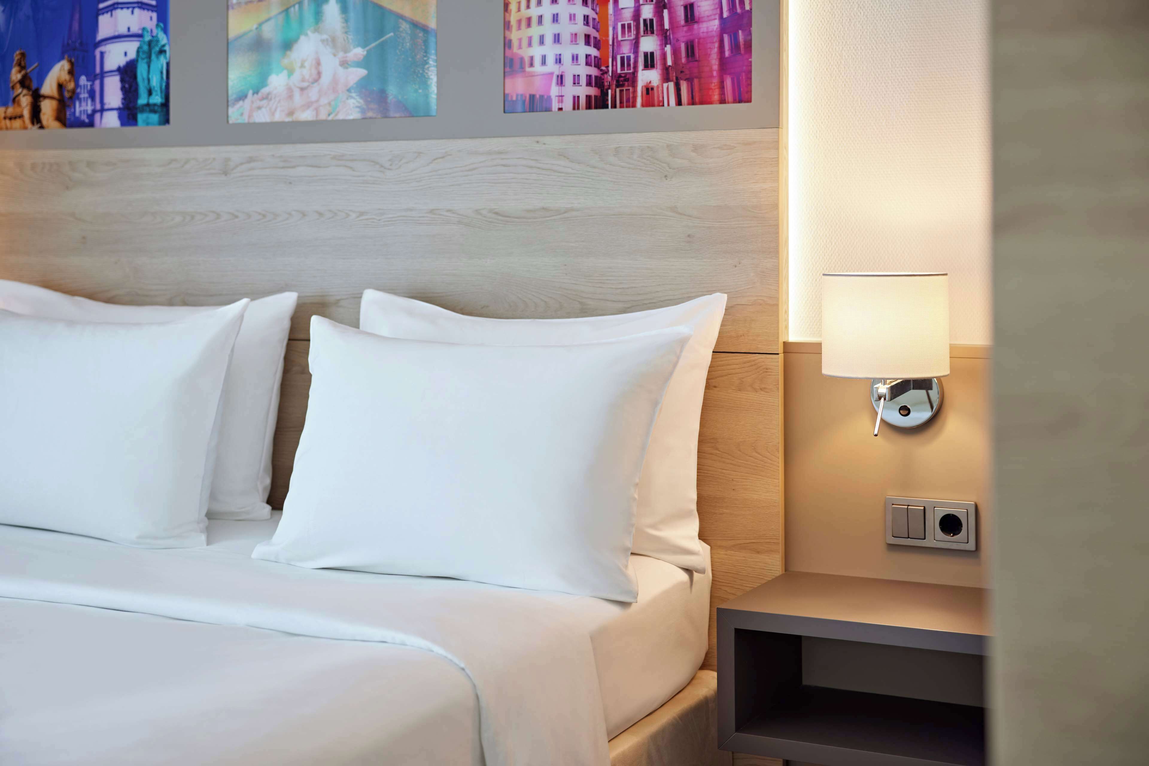 Un lit bien fait, avec une literie et des oreillers blancs, une lampe de chevet et un interrupteur fixé au mur.