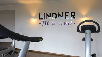 A gym with two stationary bikes, a wall with 'LINDNER - fit & well' signage, and wall-mounted lights.