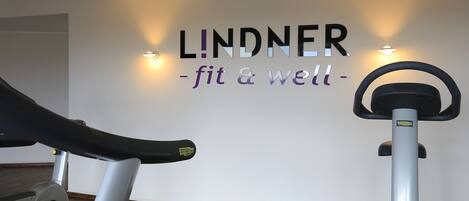 Uma academia com duas bicicletas ergométricas, uma parede com a sinalização 'LINDNER - fit & well' e luminárias fixadas na parede.