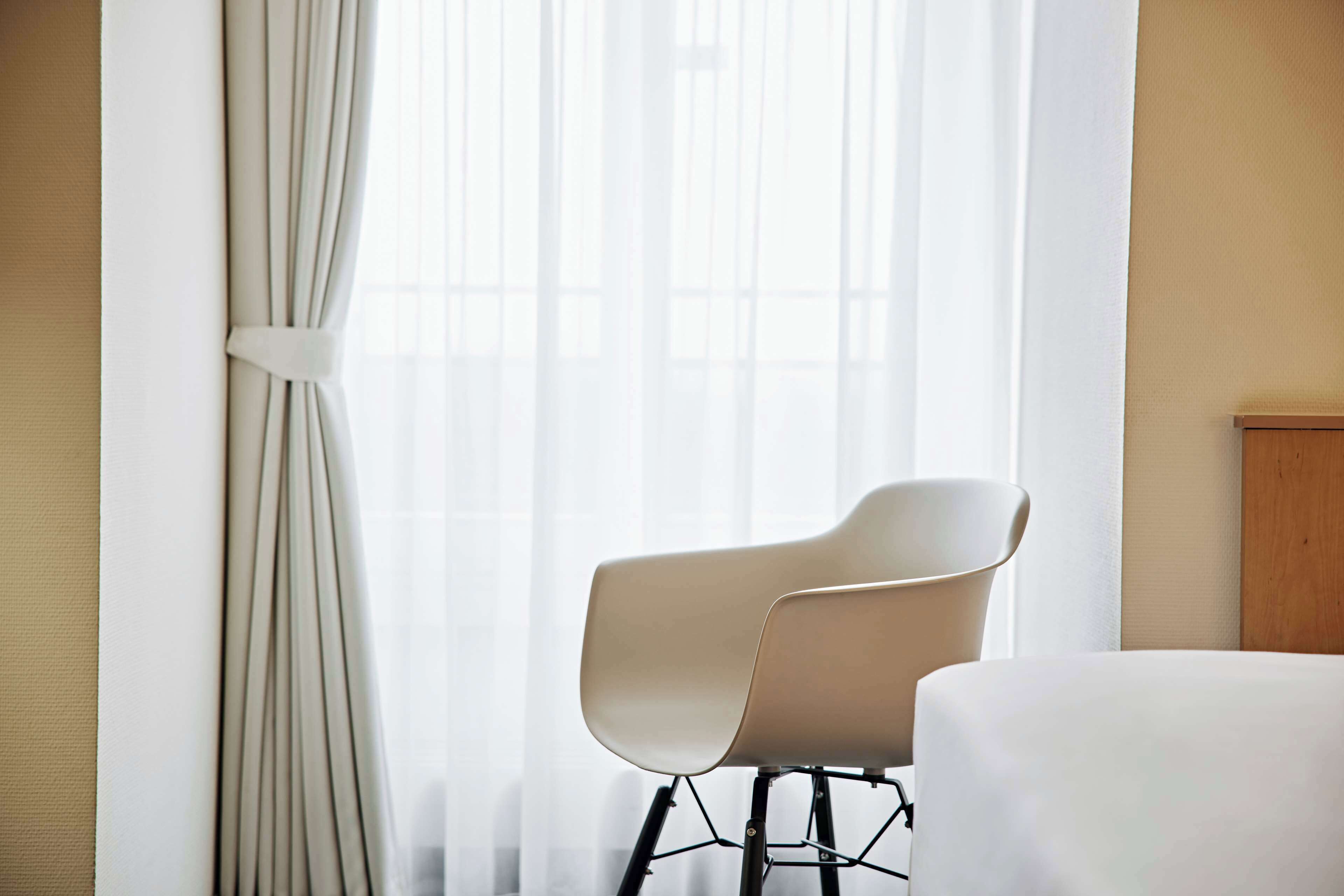 Una habitación de hotel moderna con una sola silla, una cama y una ventana con cortinas translúcidas.